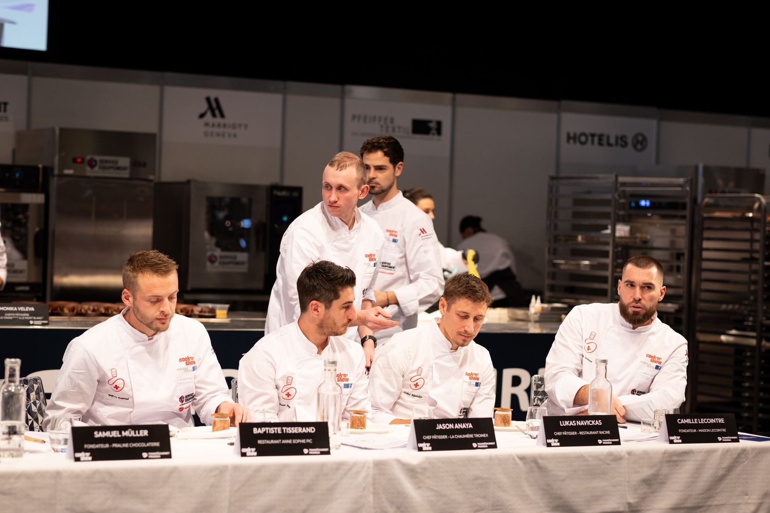 Chefs in white uniforms sit at a judging table during a professional culinary competition in a modern kitchen venue.