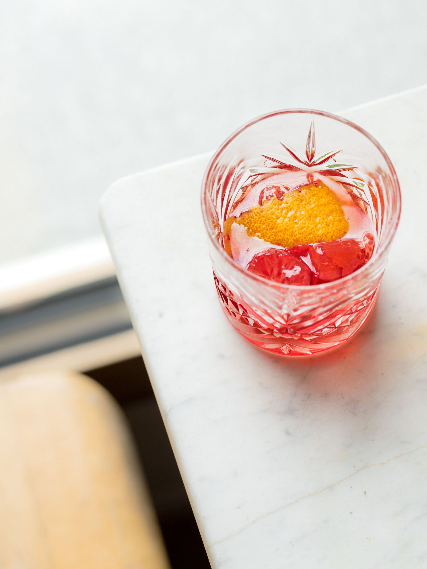 A crystal rocks glass with a pink cocktail garnished with an orange peel and cherries on a marble surface.