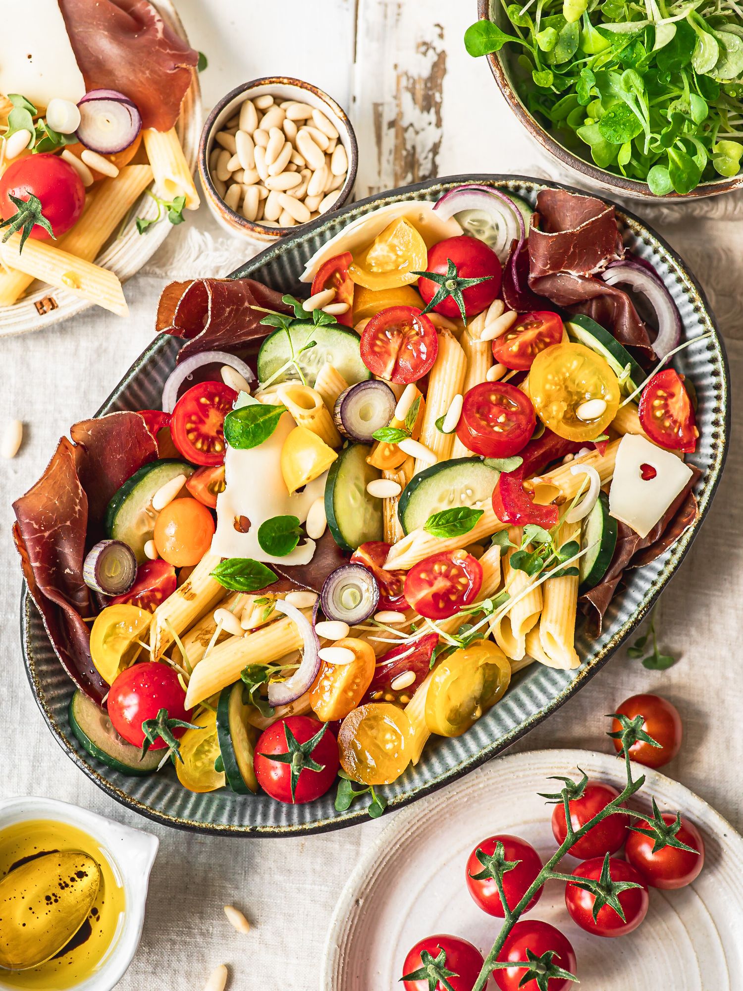 Colorful pasta salad with cherry tomatoes, zucchini, cured meat, and pine nuts on a ceramic platter.