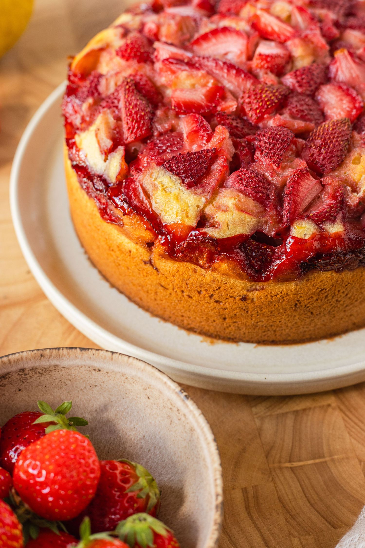 Golden strawberry upside-down cake topped with fresh glazed strawberries on white plate, with bowl of fresh strawberries beside it.