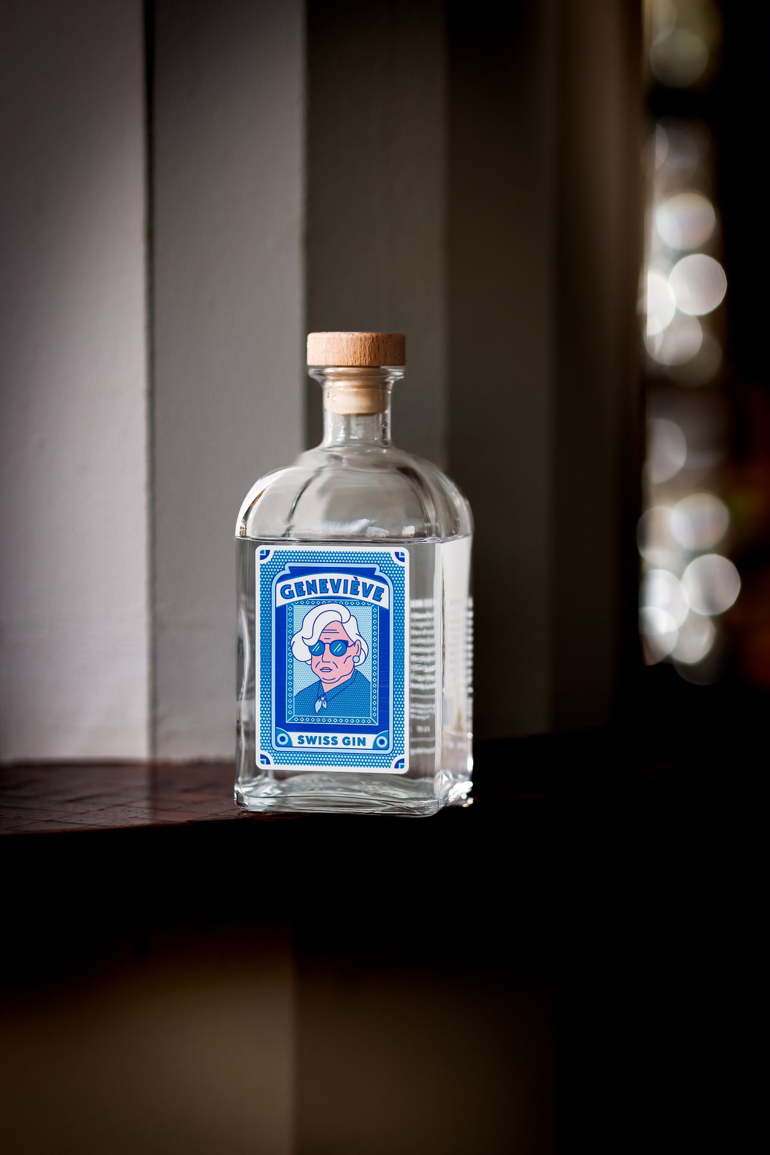 Clear glass bottle of Geneviève Swiss Gin with blue label featuring a portrait, wooden cap, on a wooden surface.