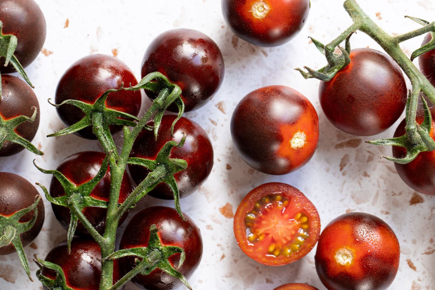 Dark purple cherry tomatoes on vine, one halved showing red interior, arranged on white surface.