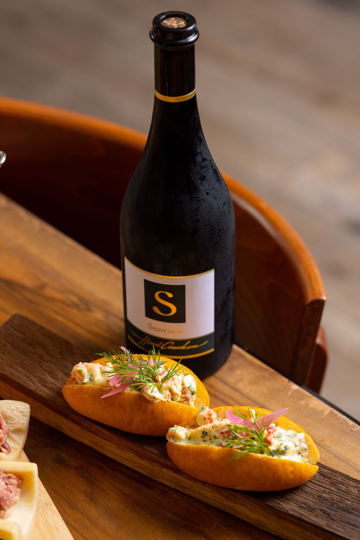 A bottle of S wine on a wooden board with two lobster rolls garnished with dill and pink flowers.