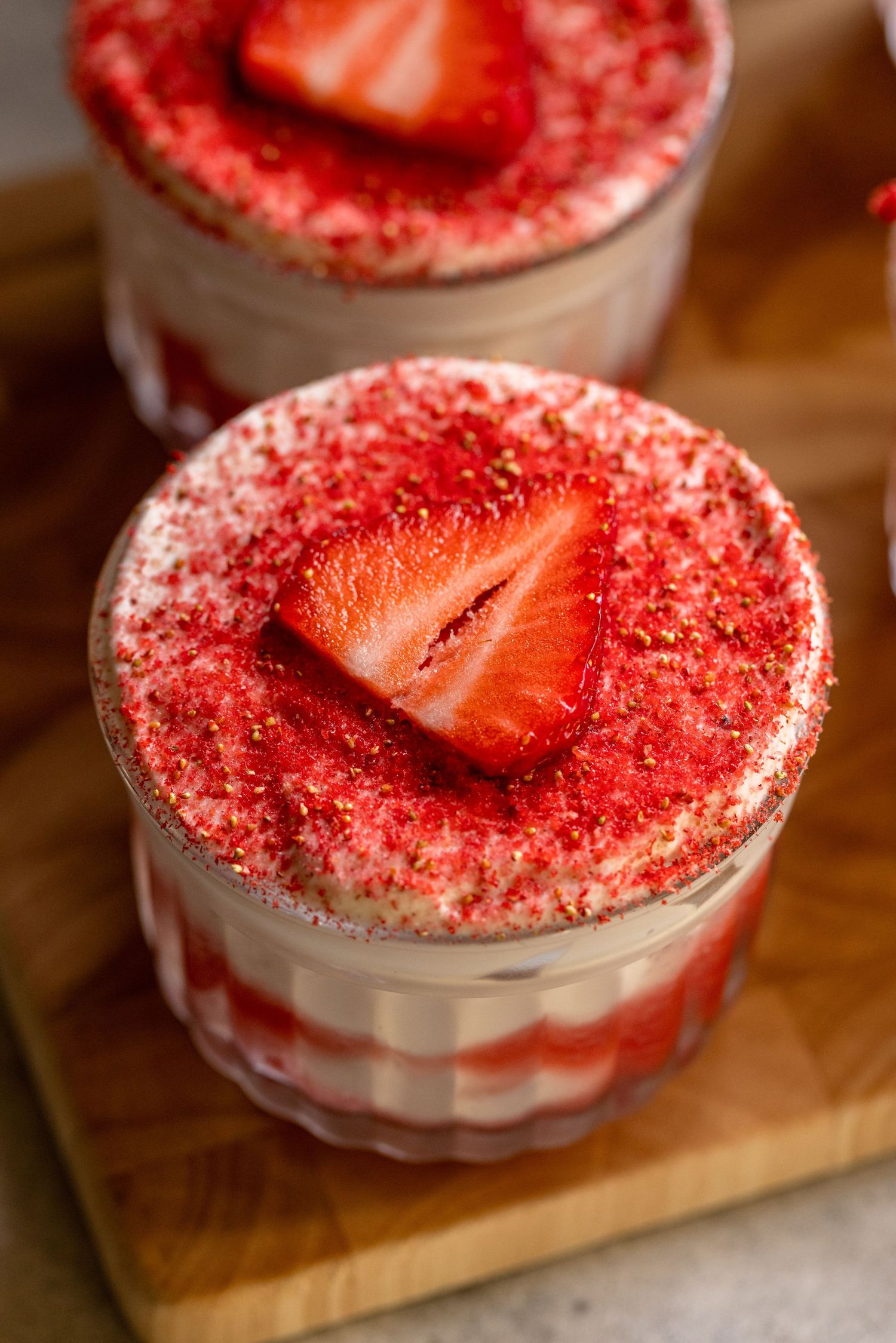 Two elegant strawberry tiramisu desserts in glass cups, topped with red crumbs and fresh strawberry slices on a wooden board.