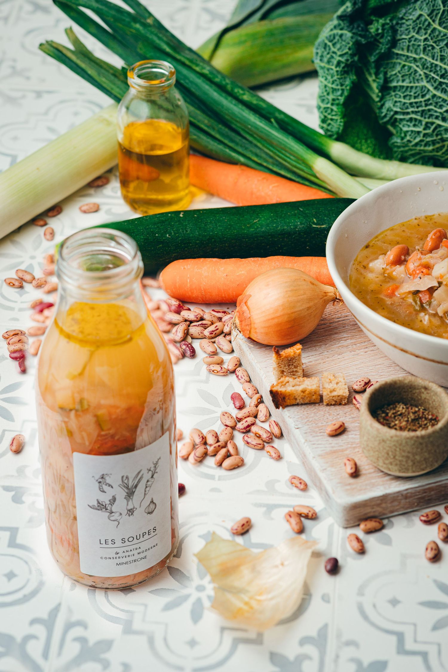 Les Soupes bottled vegetable soup with fresh leeks, carrots, onion, zucchini, beans, and olive oil on a tiled surface.