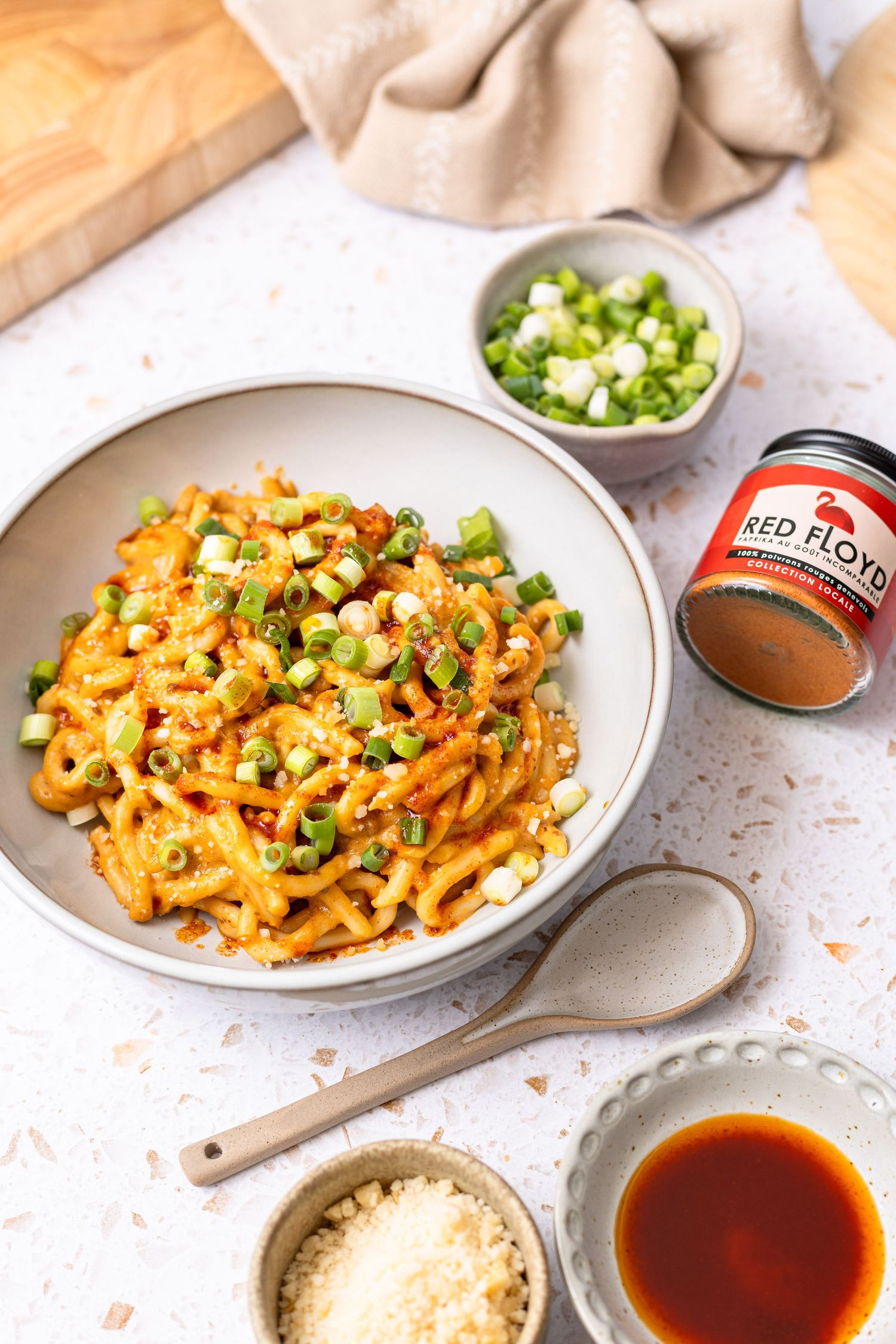 Spicy pasta dish topped with green onions in a bowl, with Red Floyd seasoning, sauce, and parmesan on white surface.