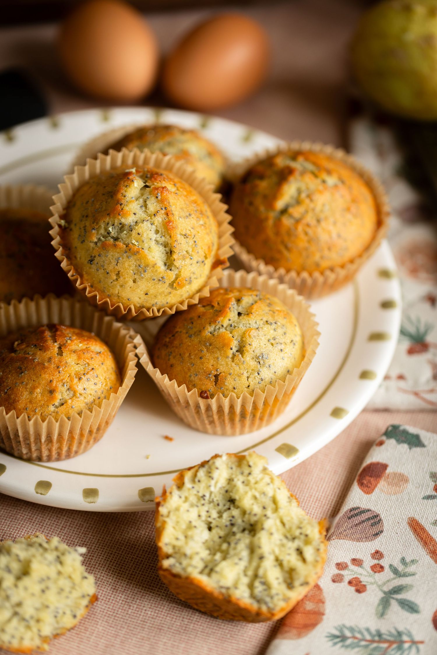 Freshly baked poppy seed muffins in paper liners on a white plate, with one muffin broken open beside brown eggs and a floral cloth.