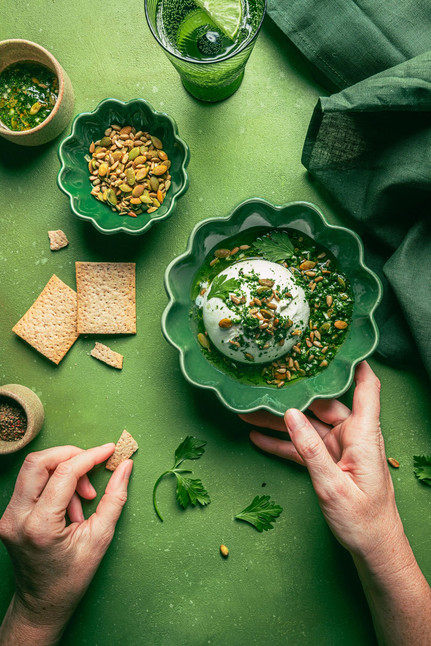 Hands holding a green bowl with burrata, herbs, pistachios on a green table with crackers and smoothie.