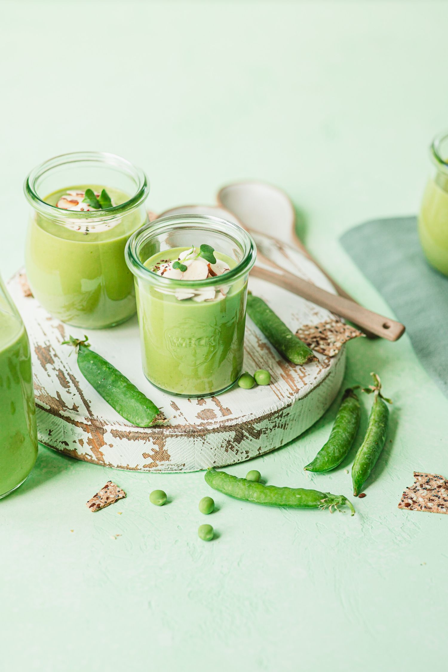 Fresh green pea soup served in glass jars with cream, garnished with microgreens, alongside pea pods on a rustic white board.