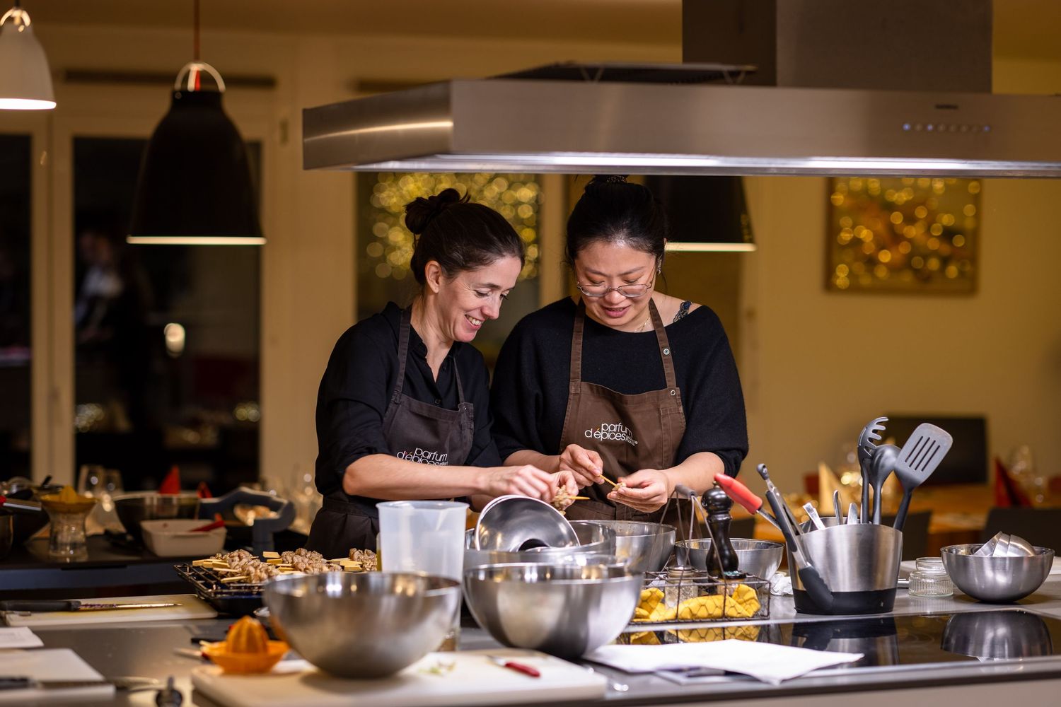 Two women in aprons cooking together in a modern kitchen, smiling while preparing food at a well-equipped cooking station.