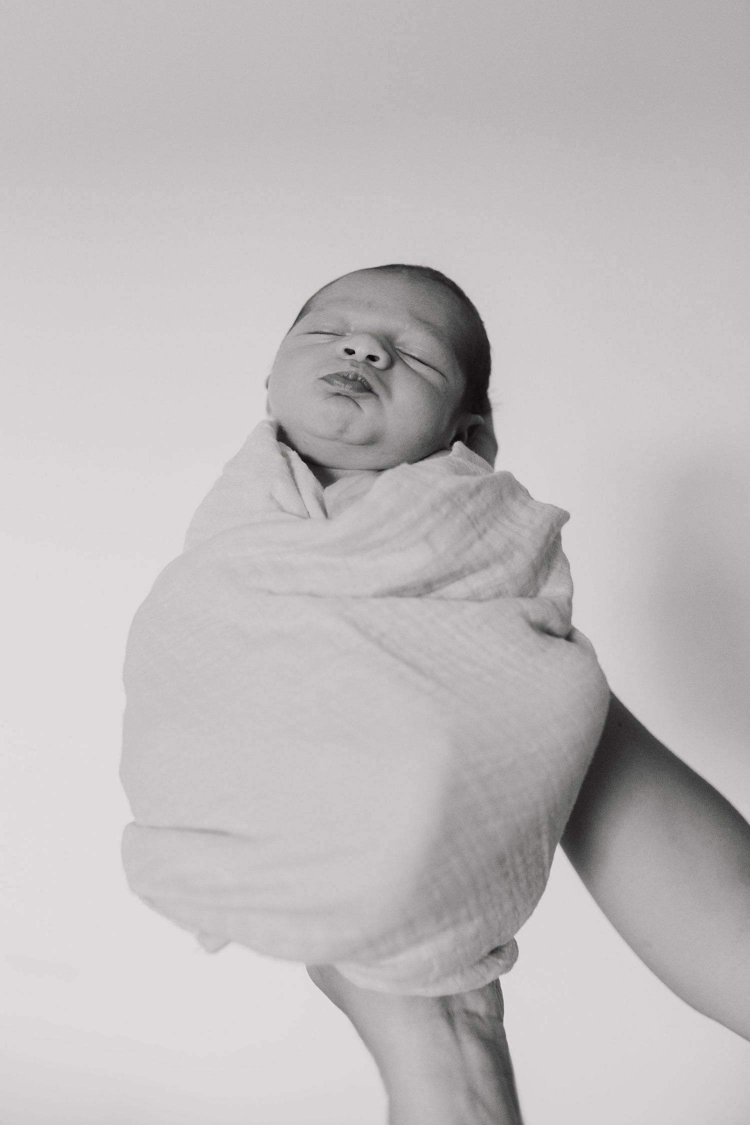 A peaceful newborn baby swaddled in white cloth is held up against a light background in black and white photography.