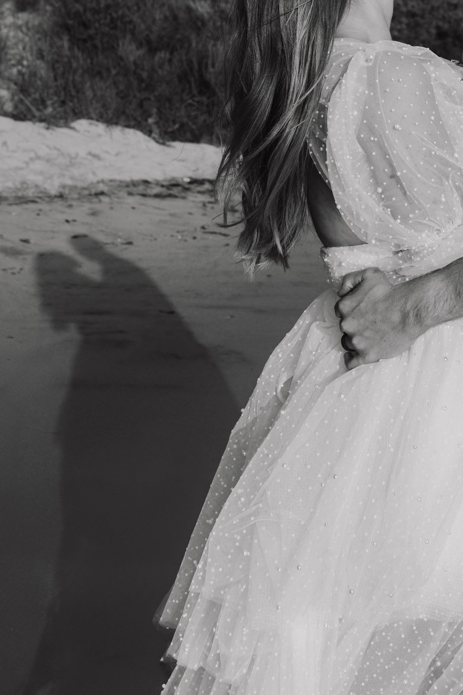 A person in a flowing white lace dress stands on a beach in black and white photography.