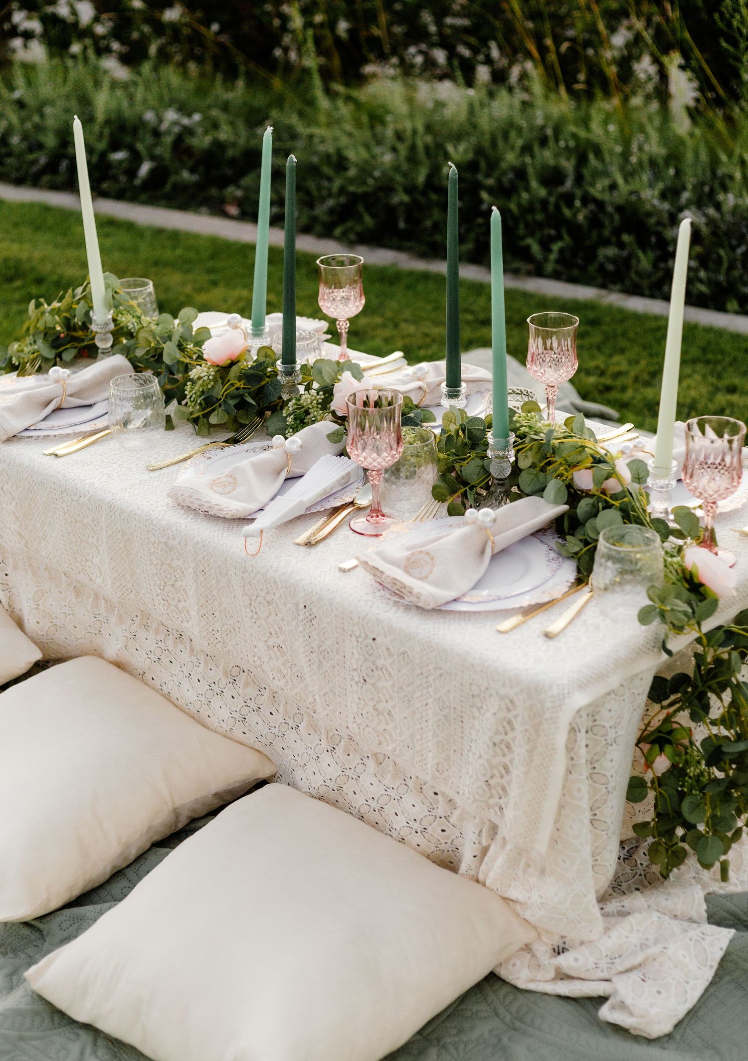 Elegant table setting with white lace tablecloth, green taper candles, and delicate floral greenery garland for outdoor dining.
