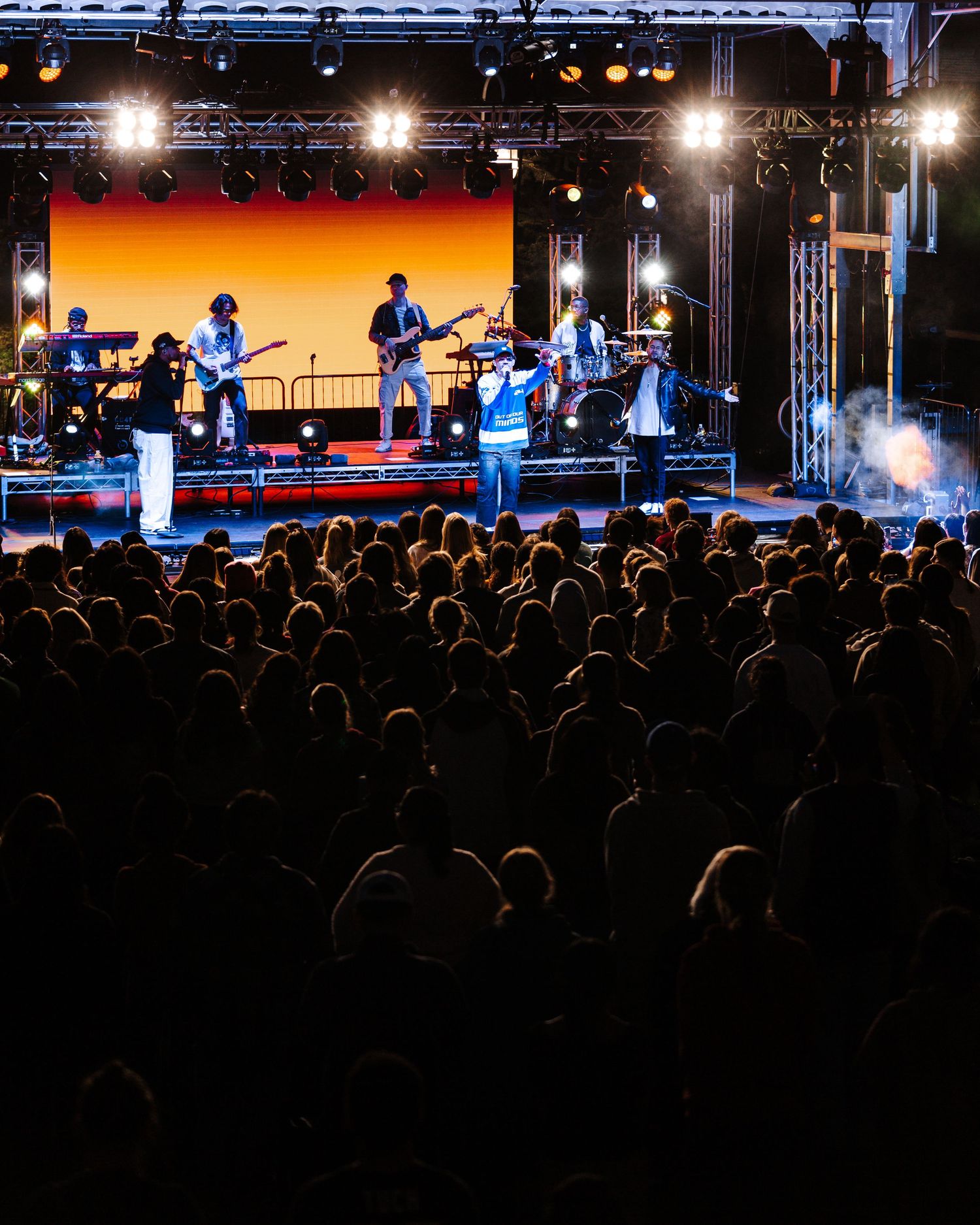 Silhouetted crowd watches performers on an illuminated stage with bright spotlights.