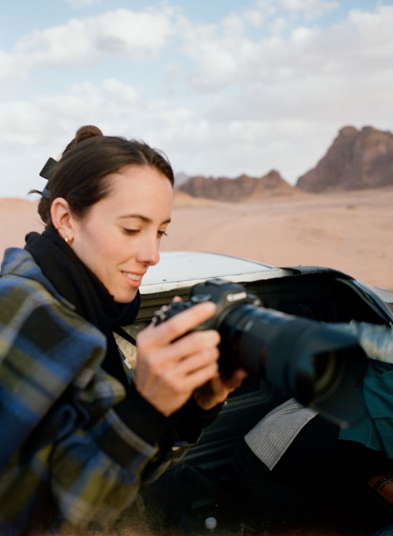 Woman in plaid jacket reviewing photos on camera in desert landscape with rocky mountains in background.