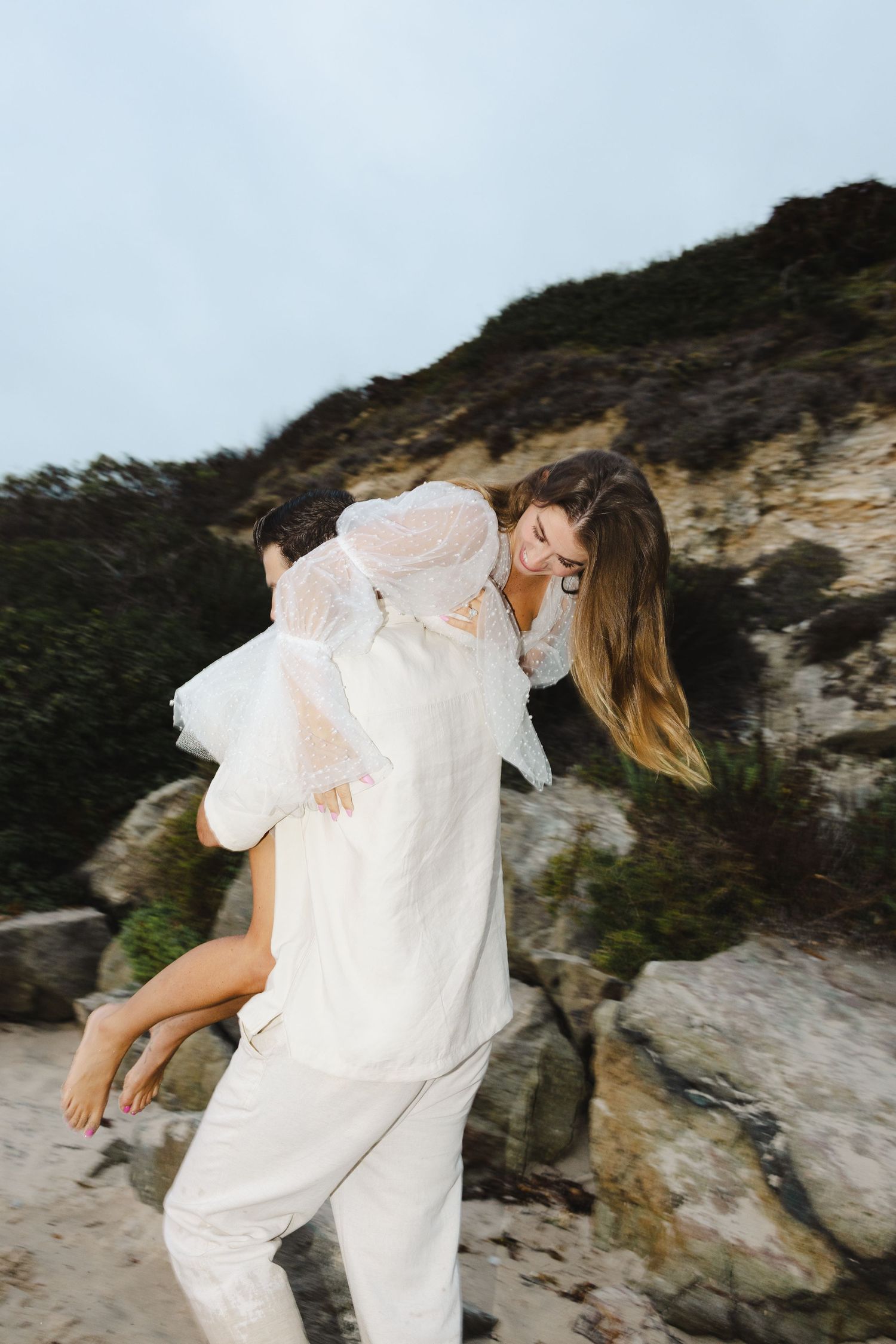 Couple in white clothing embracing on rocky coastal cliffs with wind-swept scenery in the background.
