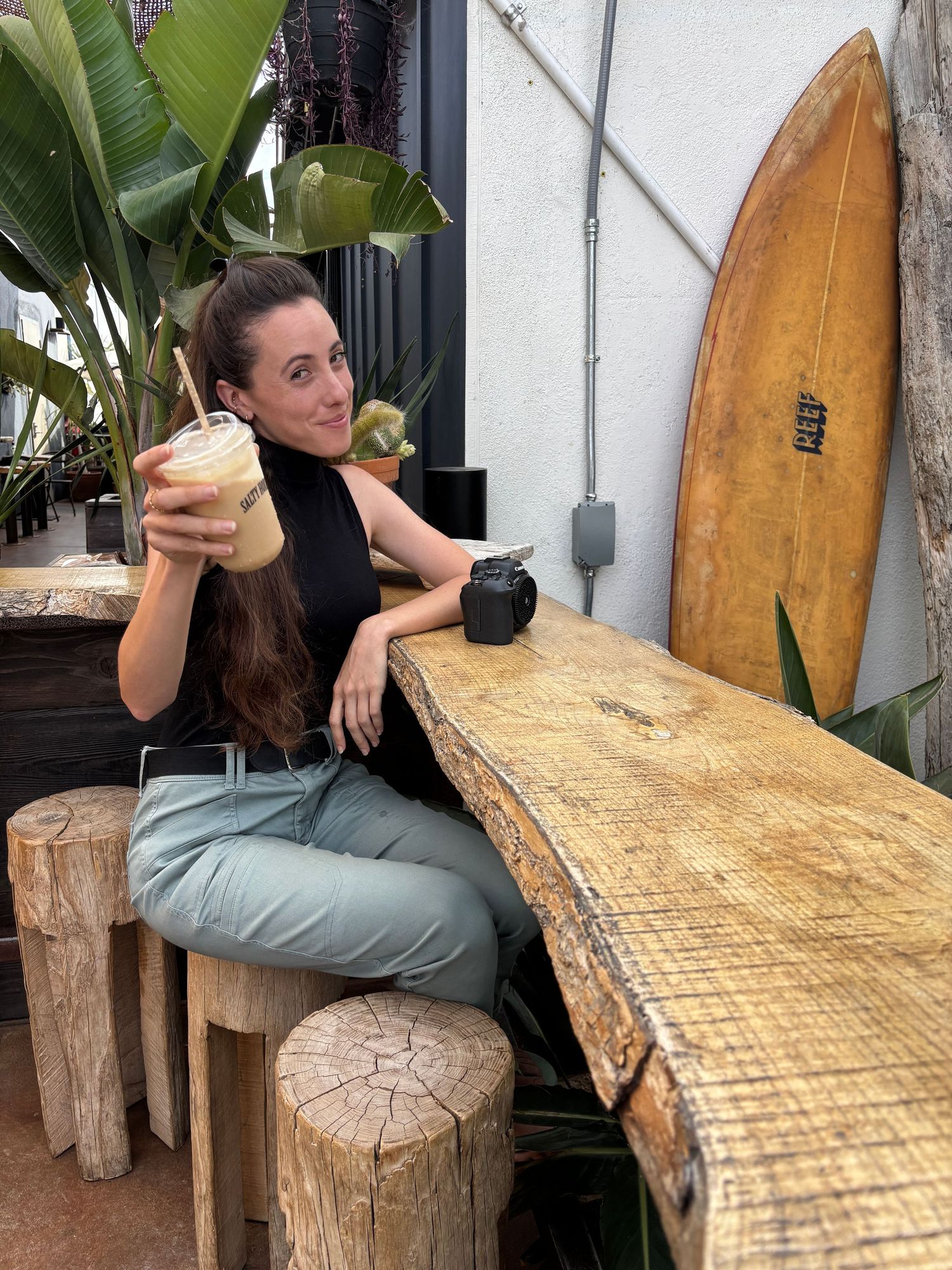 Woman holding iced coffee drink at rustic wooden bar with surfboard decor and tropical plants.
