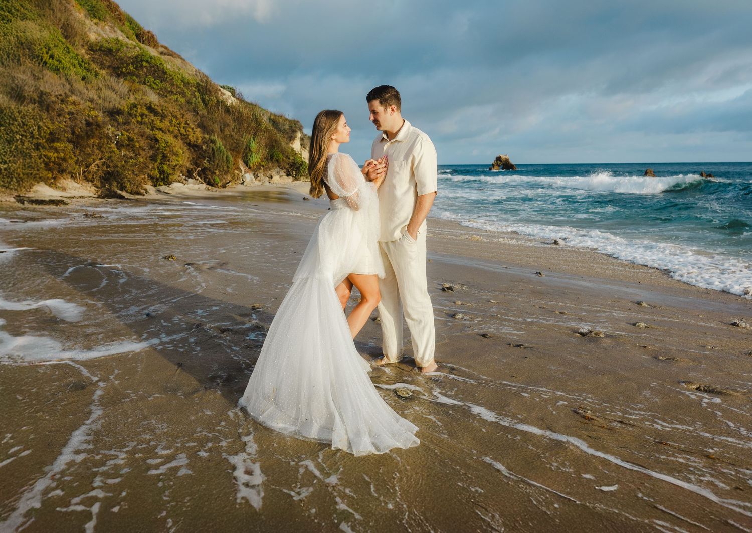 A couple in wedding attire walks along a scenic beach with dramatic cliffs and ocean waves at sunset.