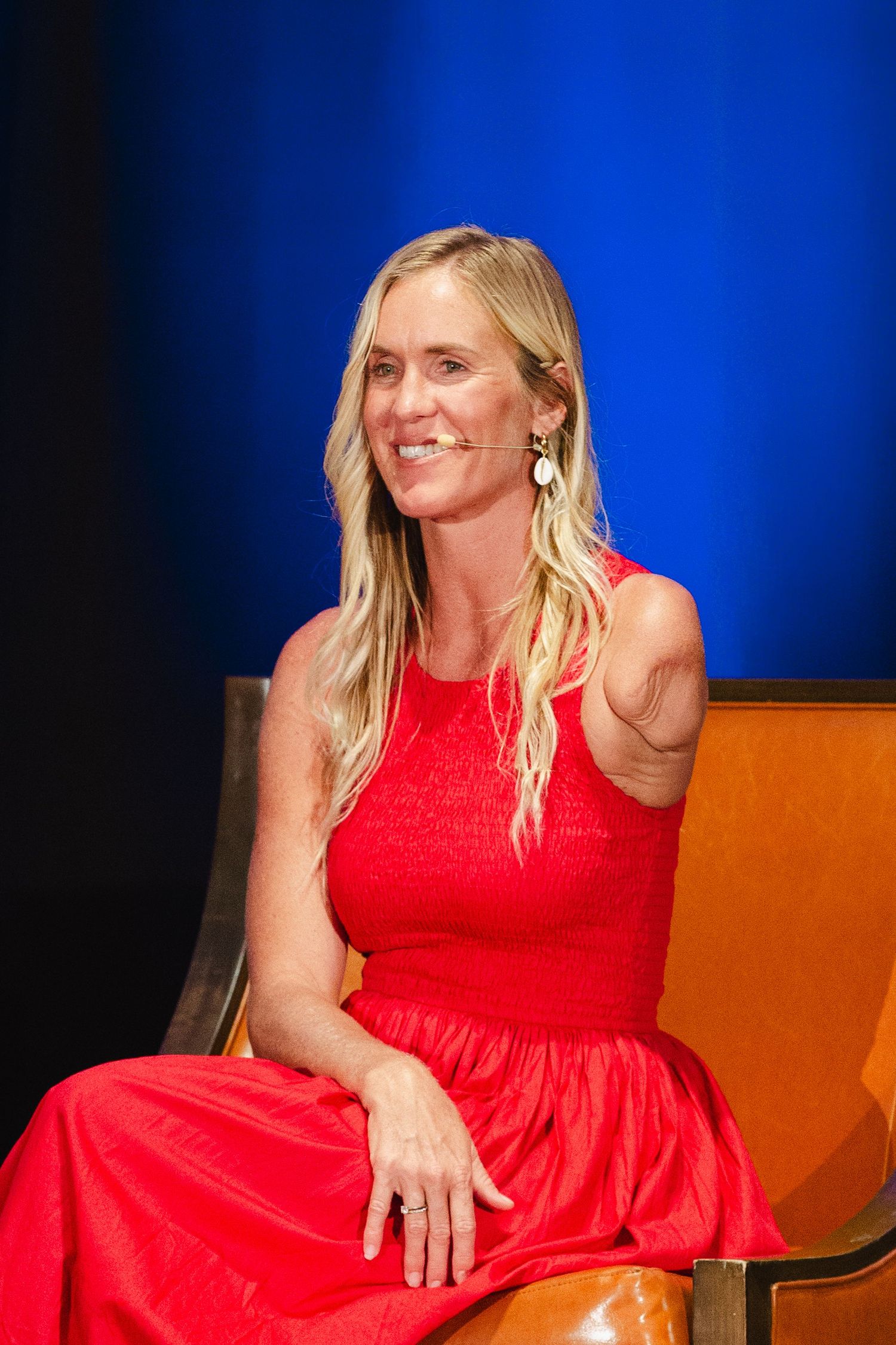 A woman in a bright red sleeveless dress sits and poses against a blue backdrop.