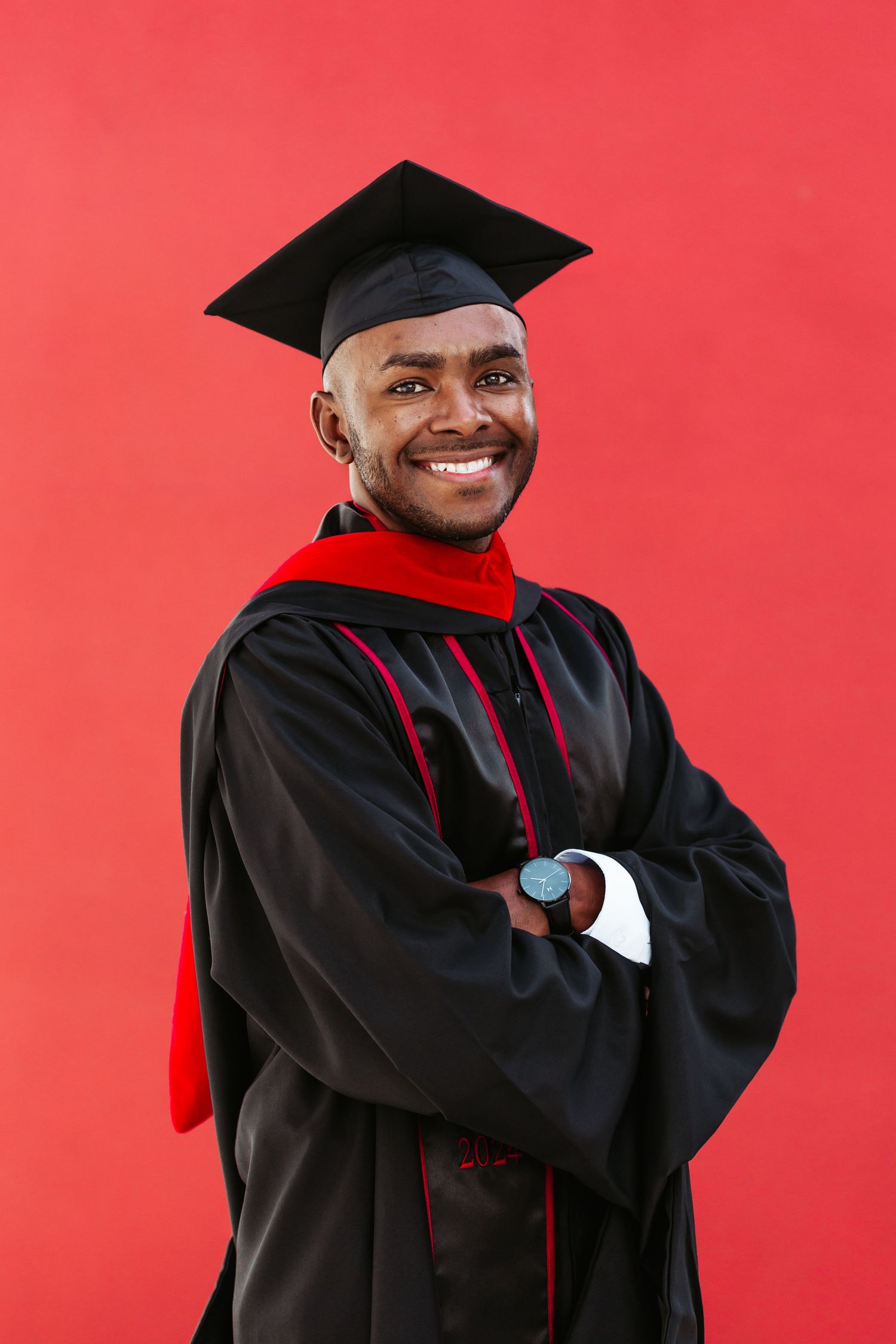 Graduate in black cap and gown with red trim smiling against a red background.