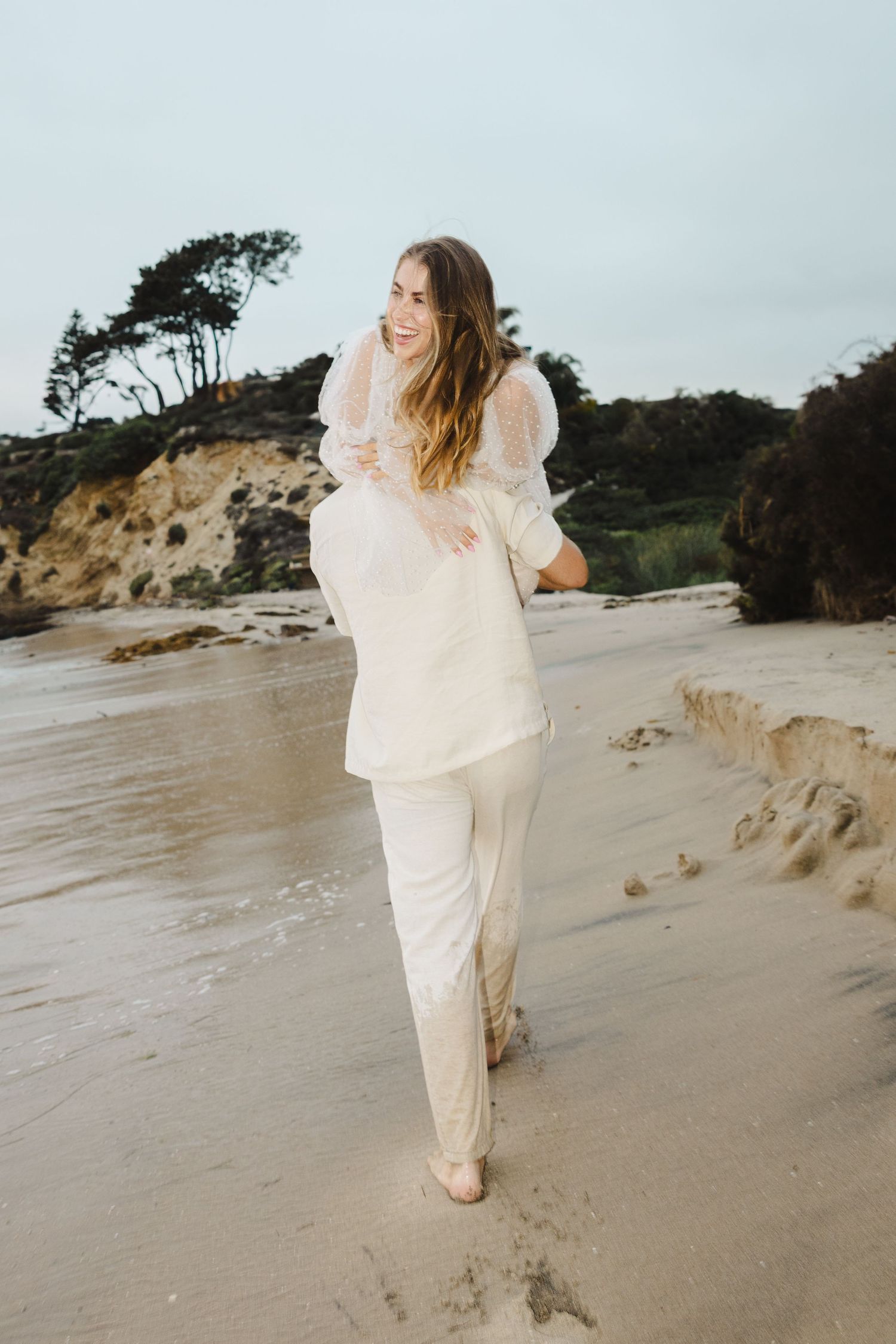 A person in flowing white clothing walks barefoot along a sandy beach near rocky cliffs at sunset.