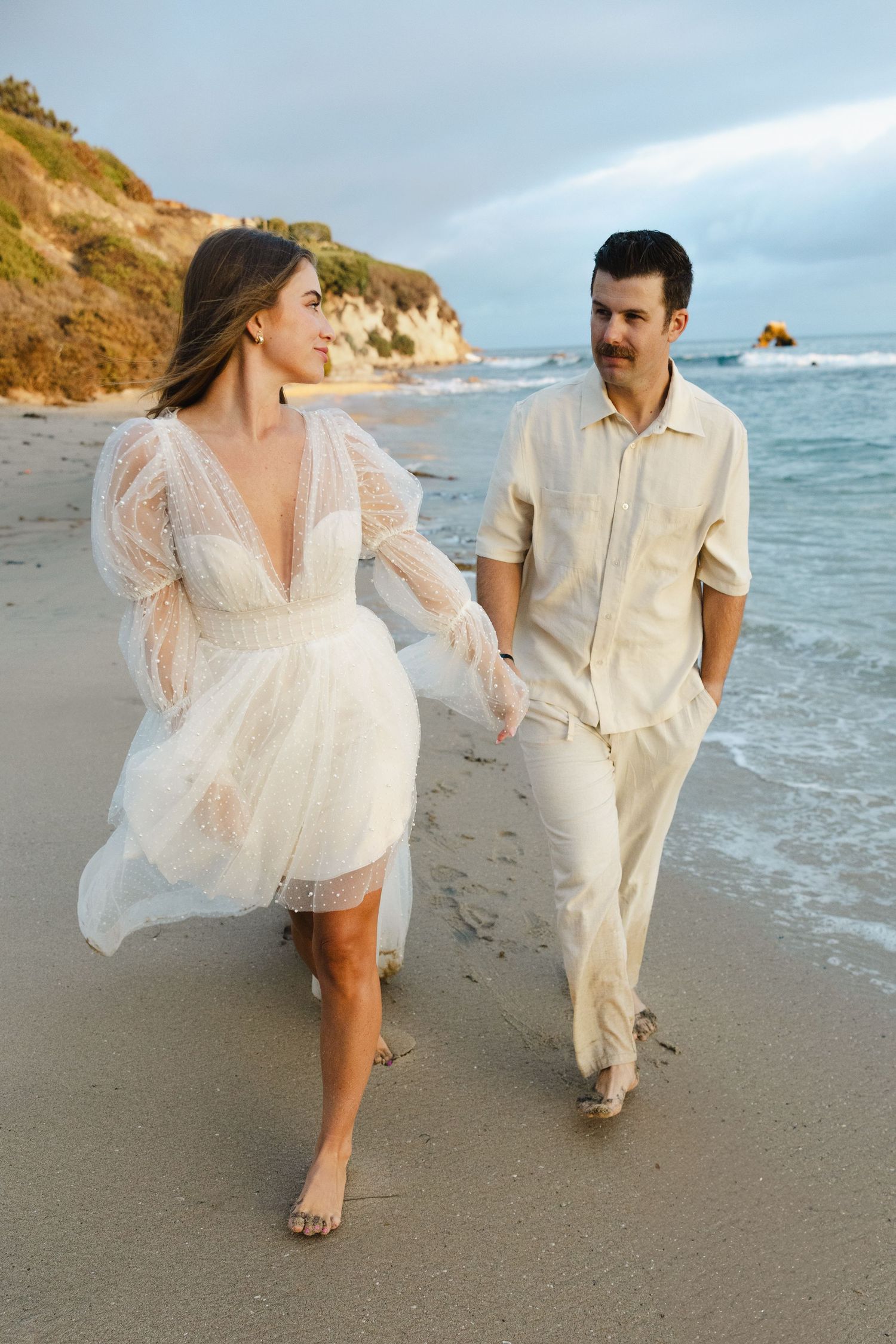 A couple in white outfits dances playfully along a scenic beach shoreline at sunset with waves crashing behind them.