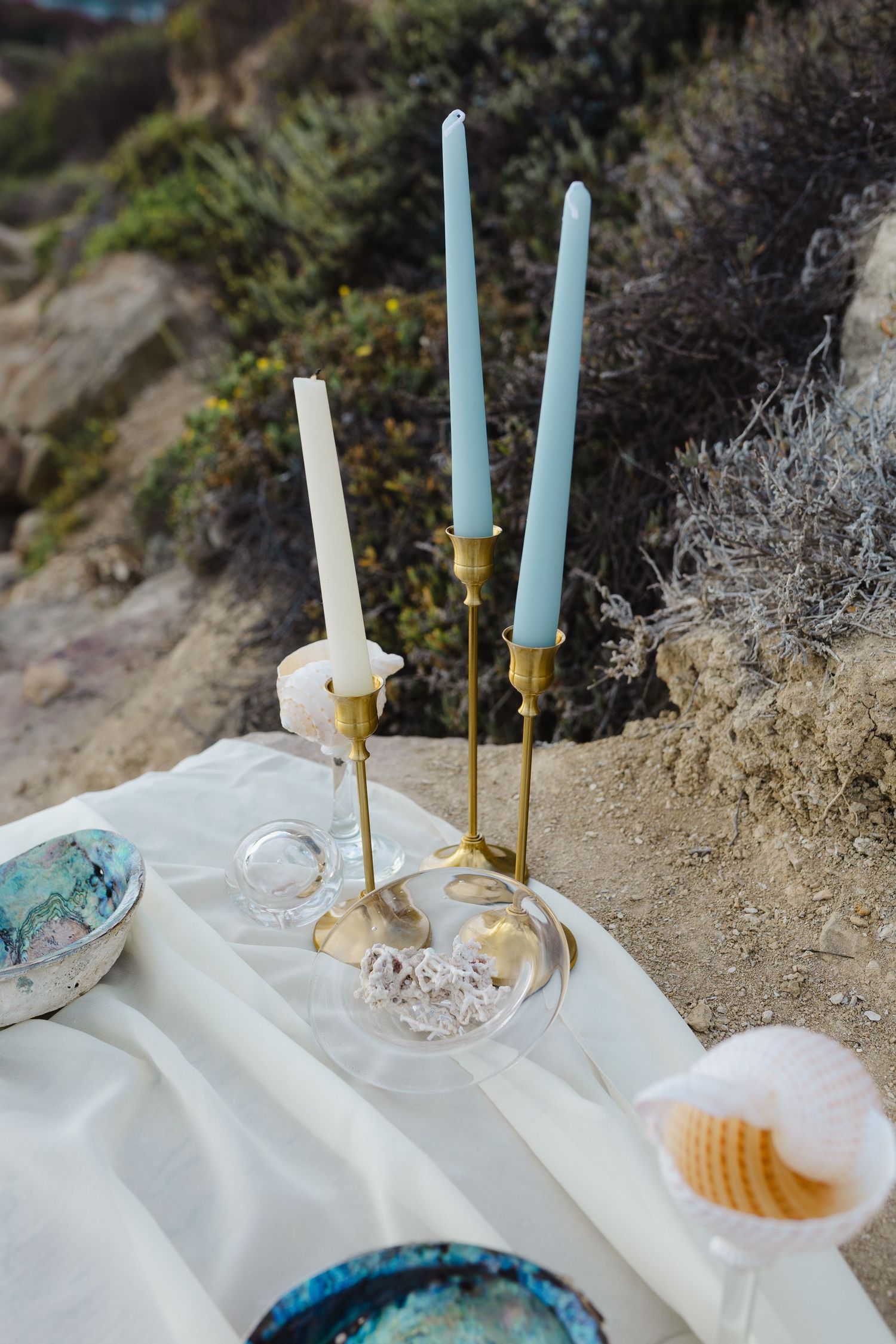 Elegant beach picnic setup with brass candlesticks, white linens, and turquoise accents against a sandy backdrop.