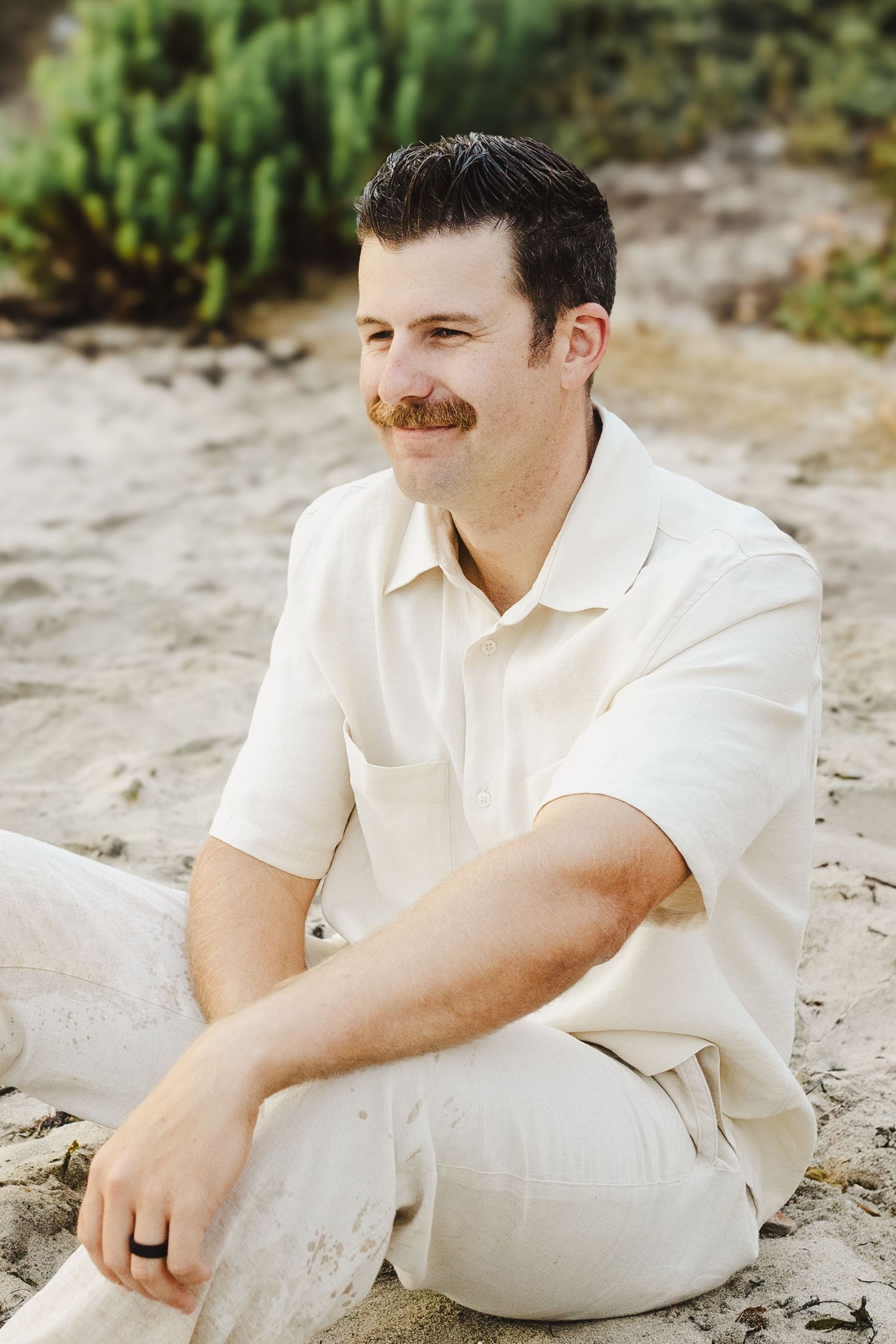 Person in a cream colored polo shirt and pants sits casually on sand with greenery in the background.