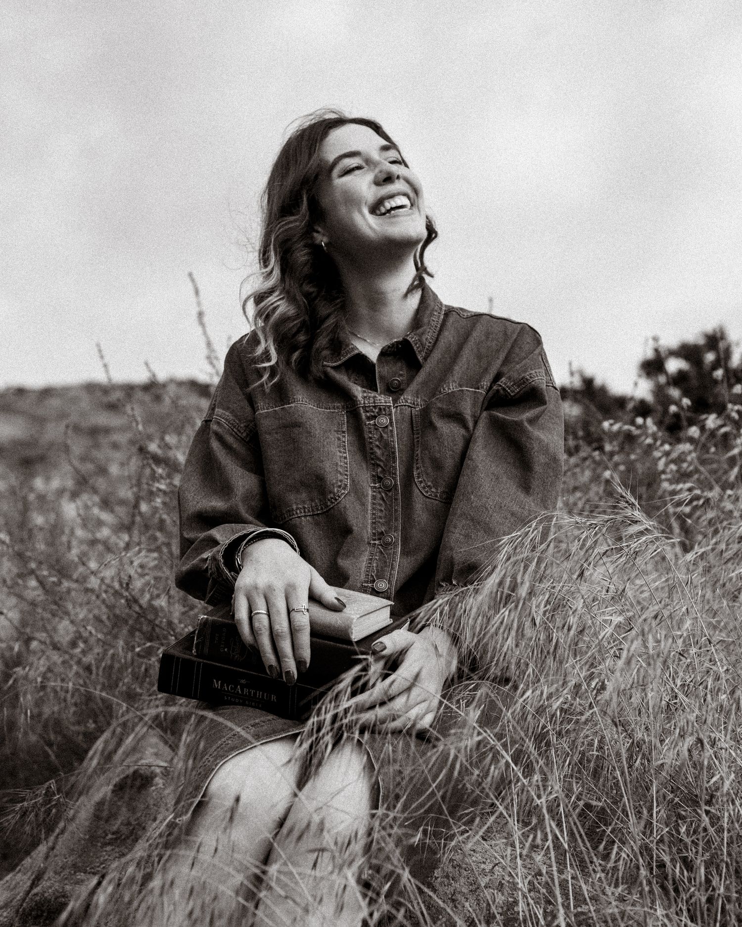 Black and white portrait of someone laughing joyfully while sitting in tall grass wearing a denim jacket.