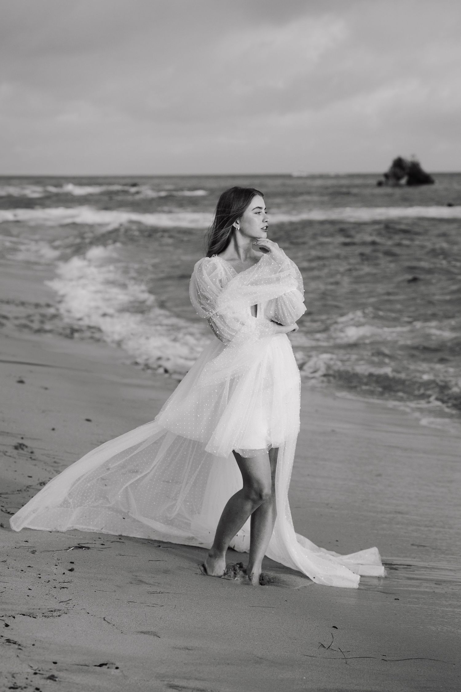 Ethereal white dress flows in the ocean breeze as figure poses gracefully on moody beach at sunset.