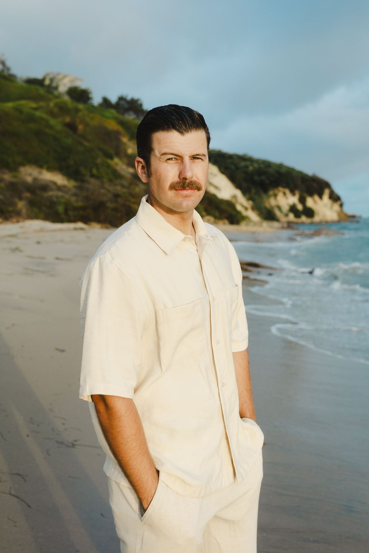 A person in a white button-down shirt stands on a scenic beach with cliffs and ocean waves in the background.