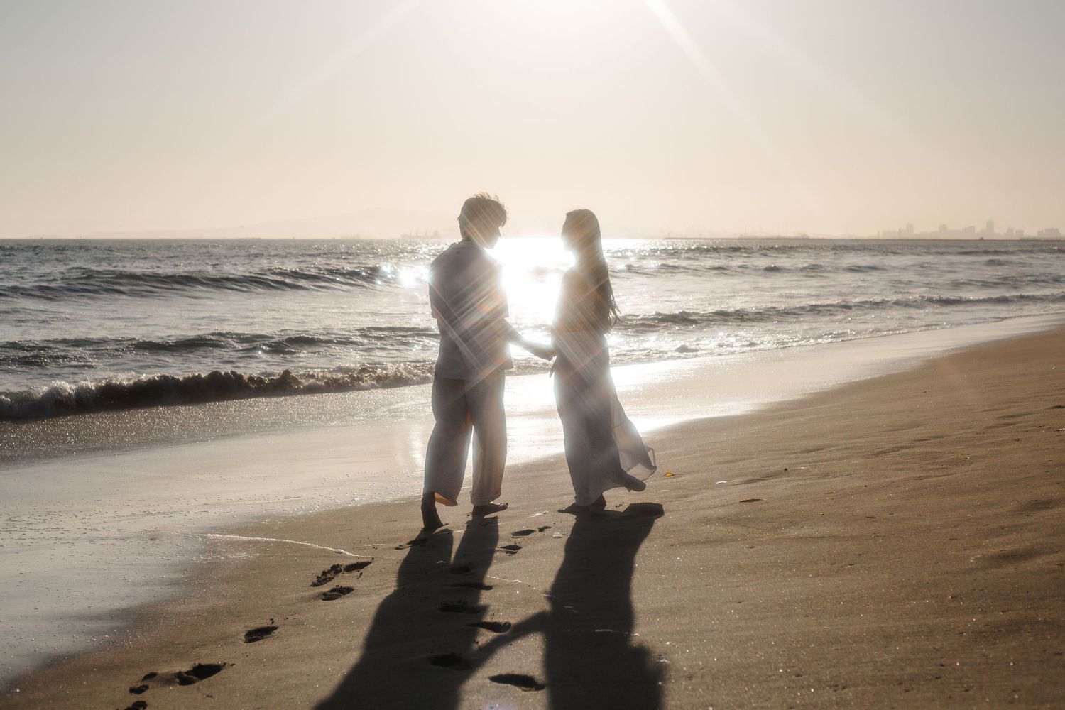 Silhouettes of couple walking along beach at sunset with dogs casting long shadows on sandy shore.