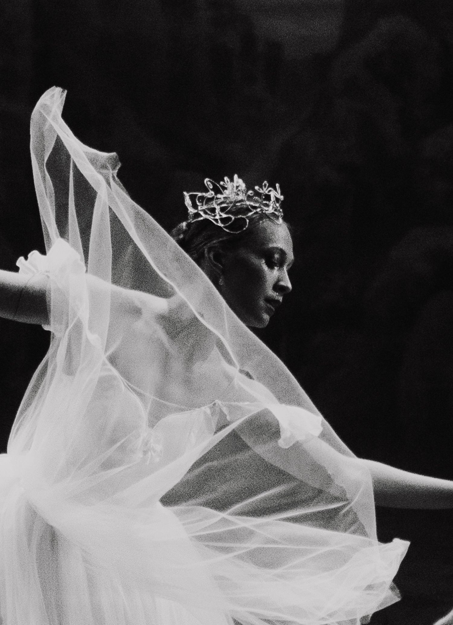 A dramatic black and white photograph of a dancer in a flowing white veil and delicate crown against a dark background.
