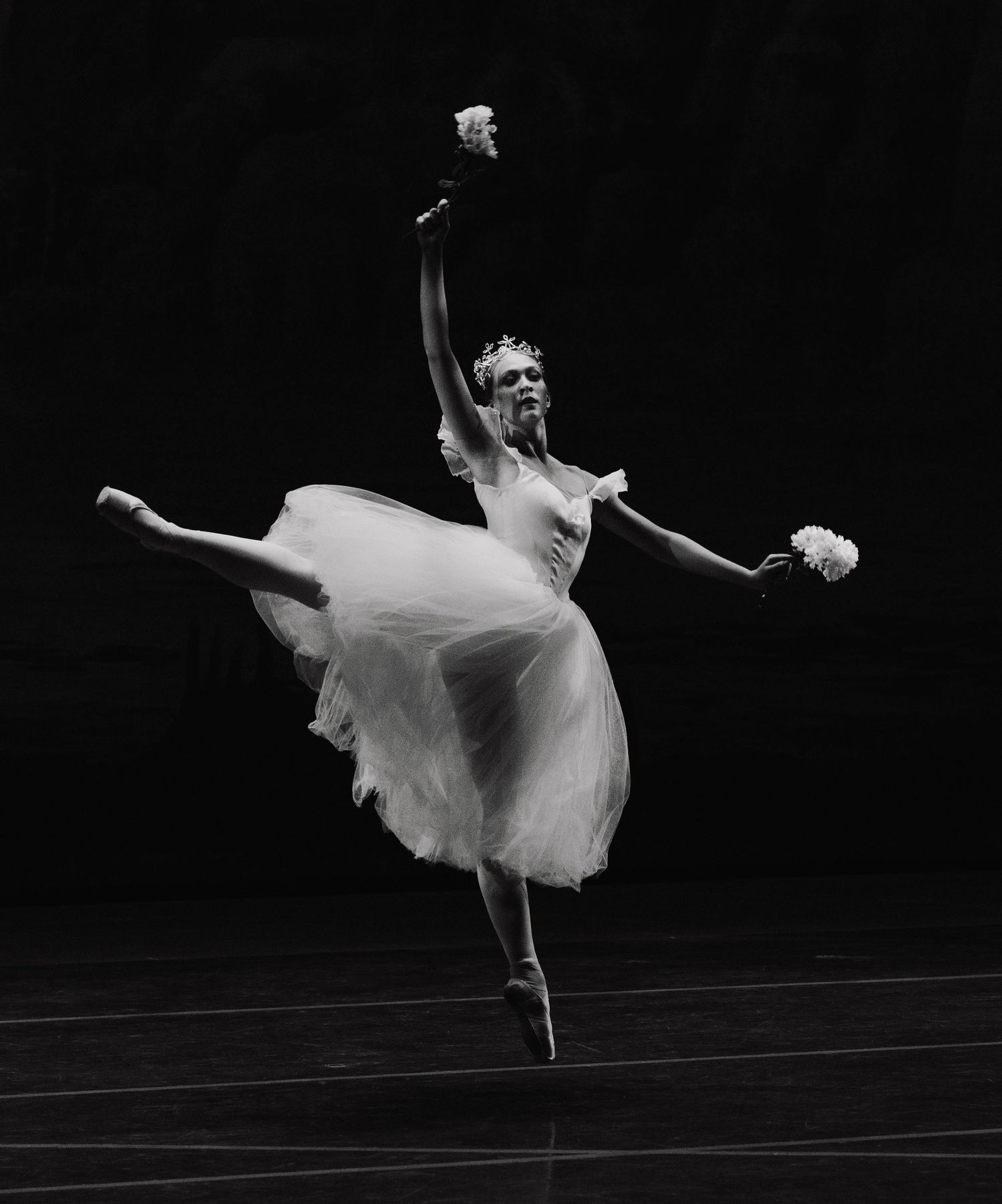 Ballet dancer in white tutu performs graceful leaps across dark stage in black and white photography.