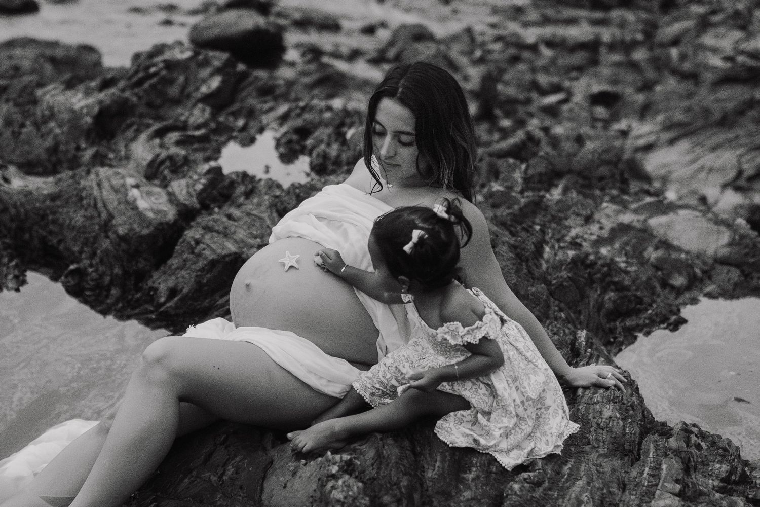 Black and white photo of a pregnant woman sitting on rocks at the beach with a young girl in a white dress.