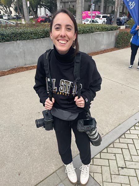 Smiling woman wearing a black Cal State sweatshirt holding two professional cameras with a telephoto lens outdoors.