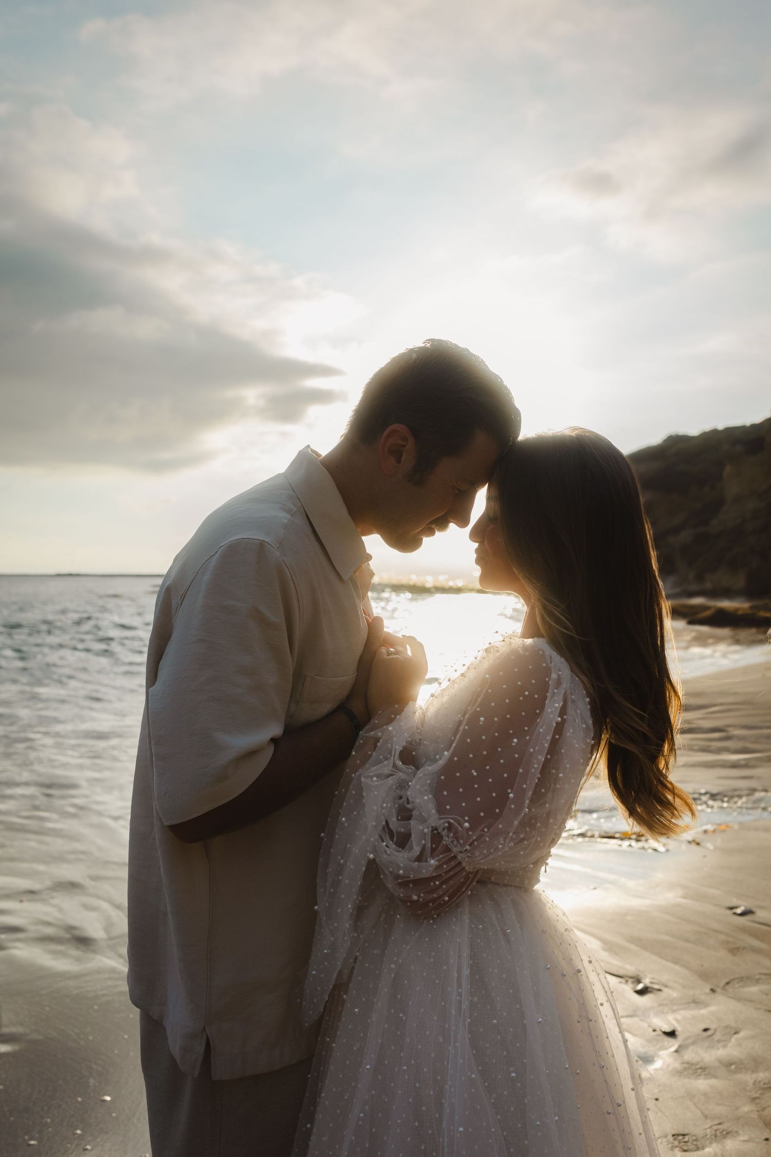 A romantic couple embraces on a sunlit beach at golden hour as waves crash on the shore in the background.