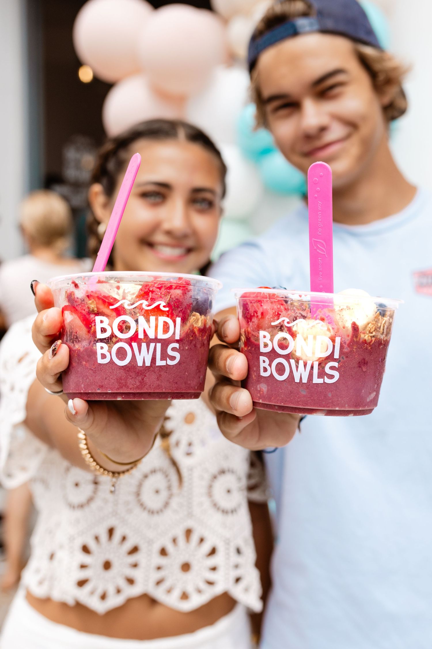 Two people holding pink smoothie bowls from Bondi Bowls with pink straws against a blurred background.