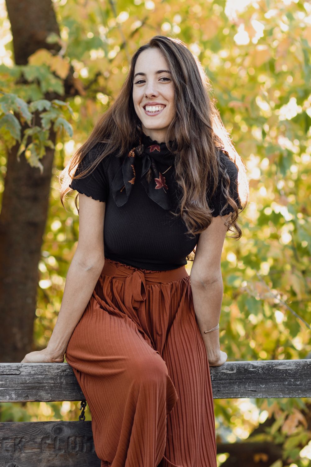 Expectant mother in a rust-colored dress and black scarf poses outdoors against autumn foliage in a photo series.