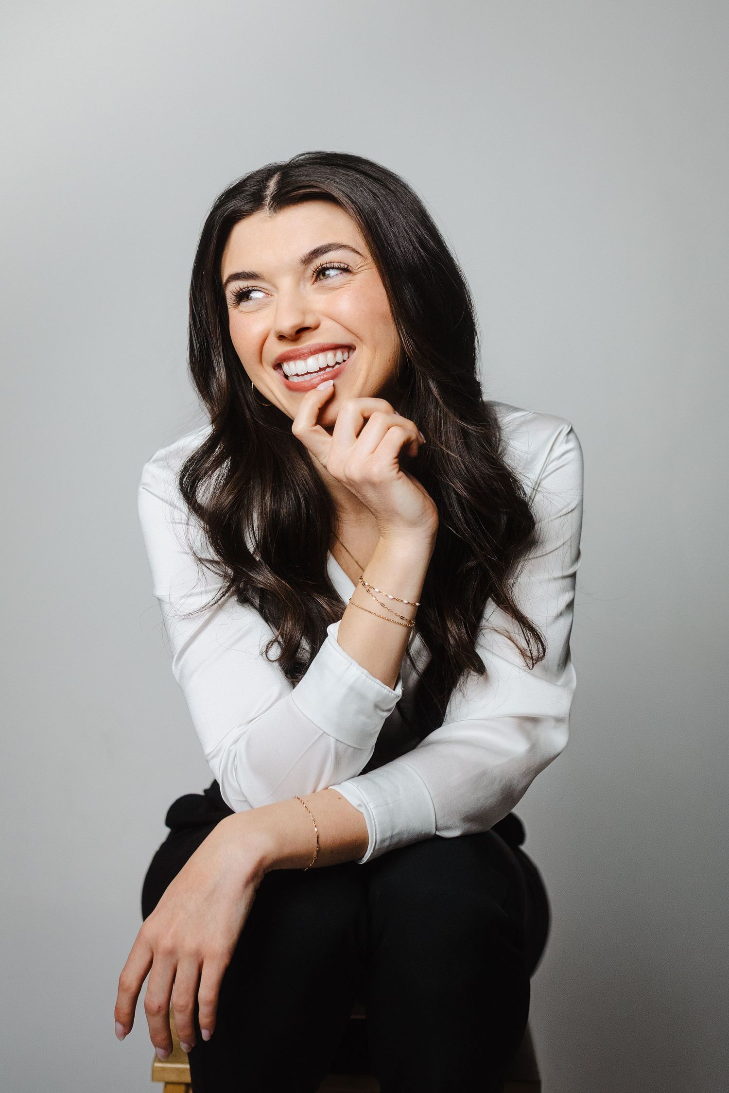Smiling woman with long dark wavy hair in a white blouse and black pants sits on a stool, hand on chin, looking away.