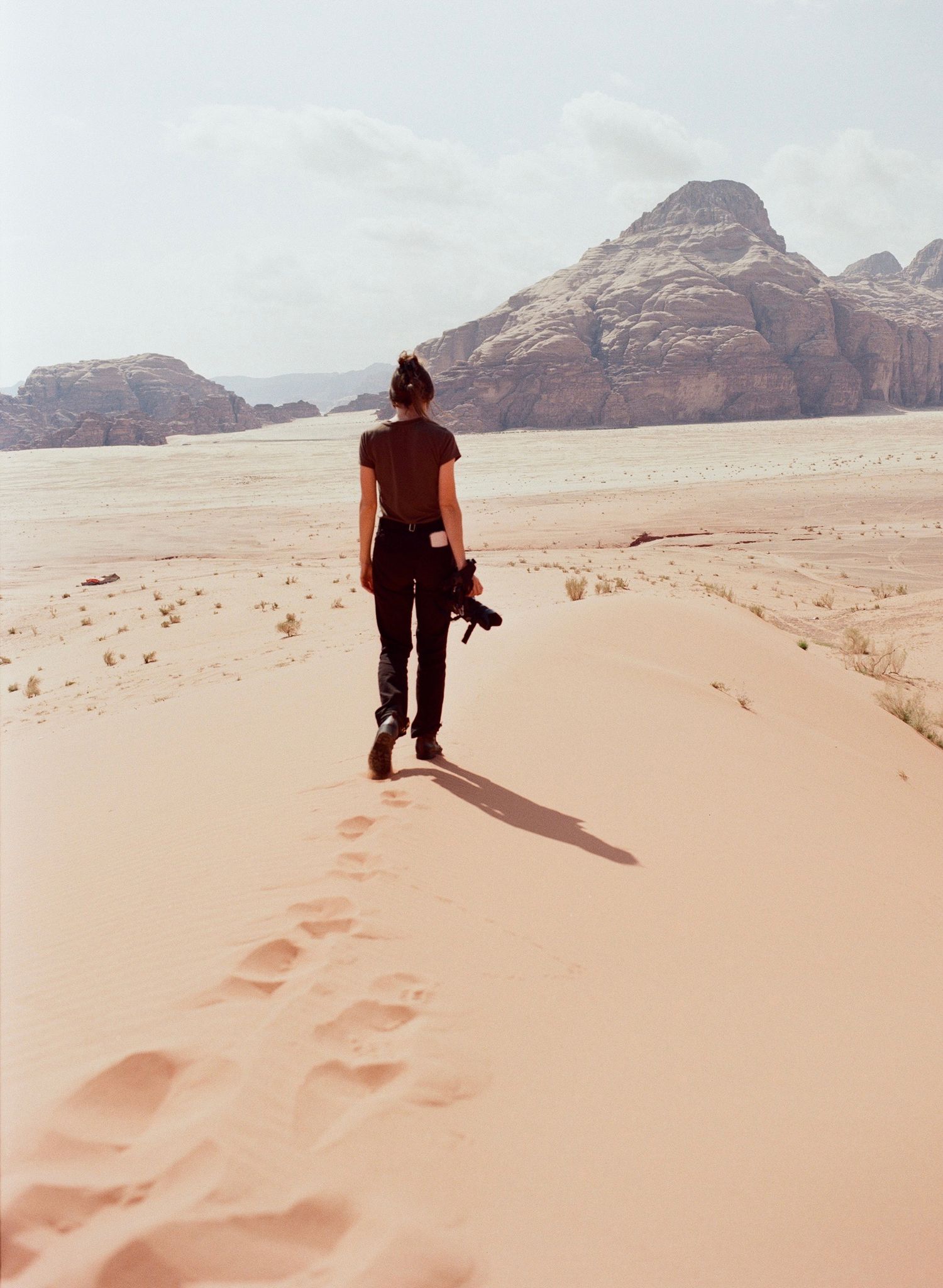 A lone photographer walks across sandy desert dunes with rocky mountains in the background, carrying a camera.