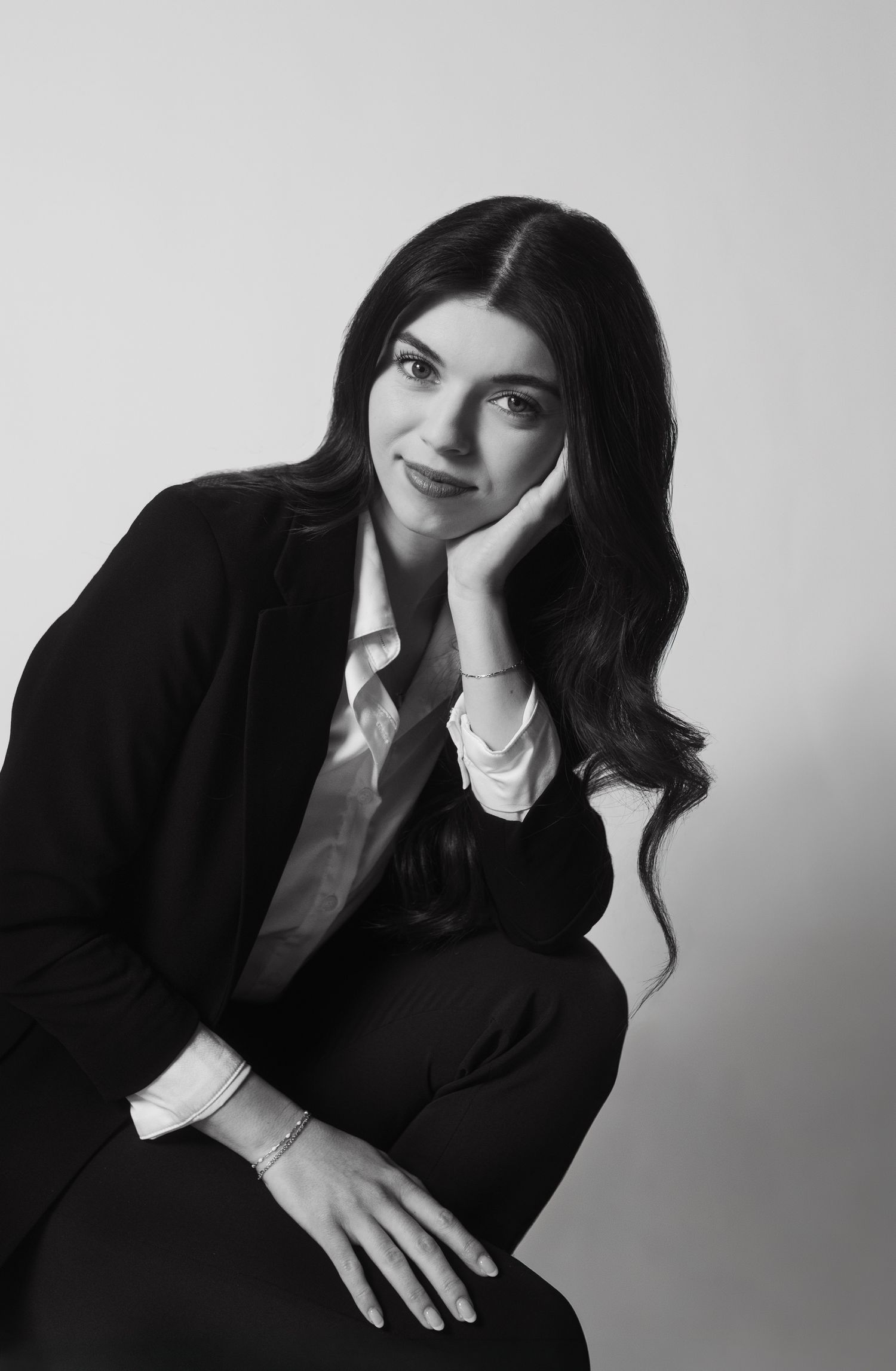 Black and white portrait of a smiling woman with long wavy hair, wearing a dark blazer and white shirt, resting her cheek on her hand.