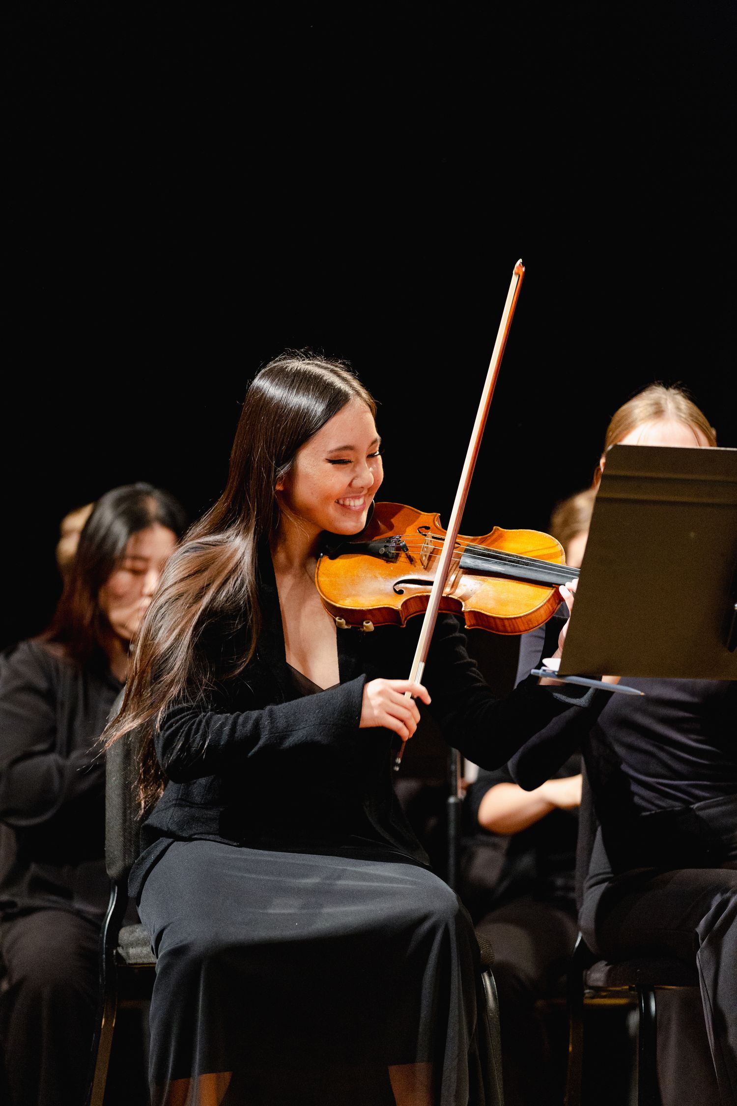 Orchestra musicians in black attire performing with violins on a dimly lit stage.