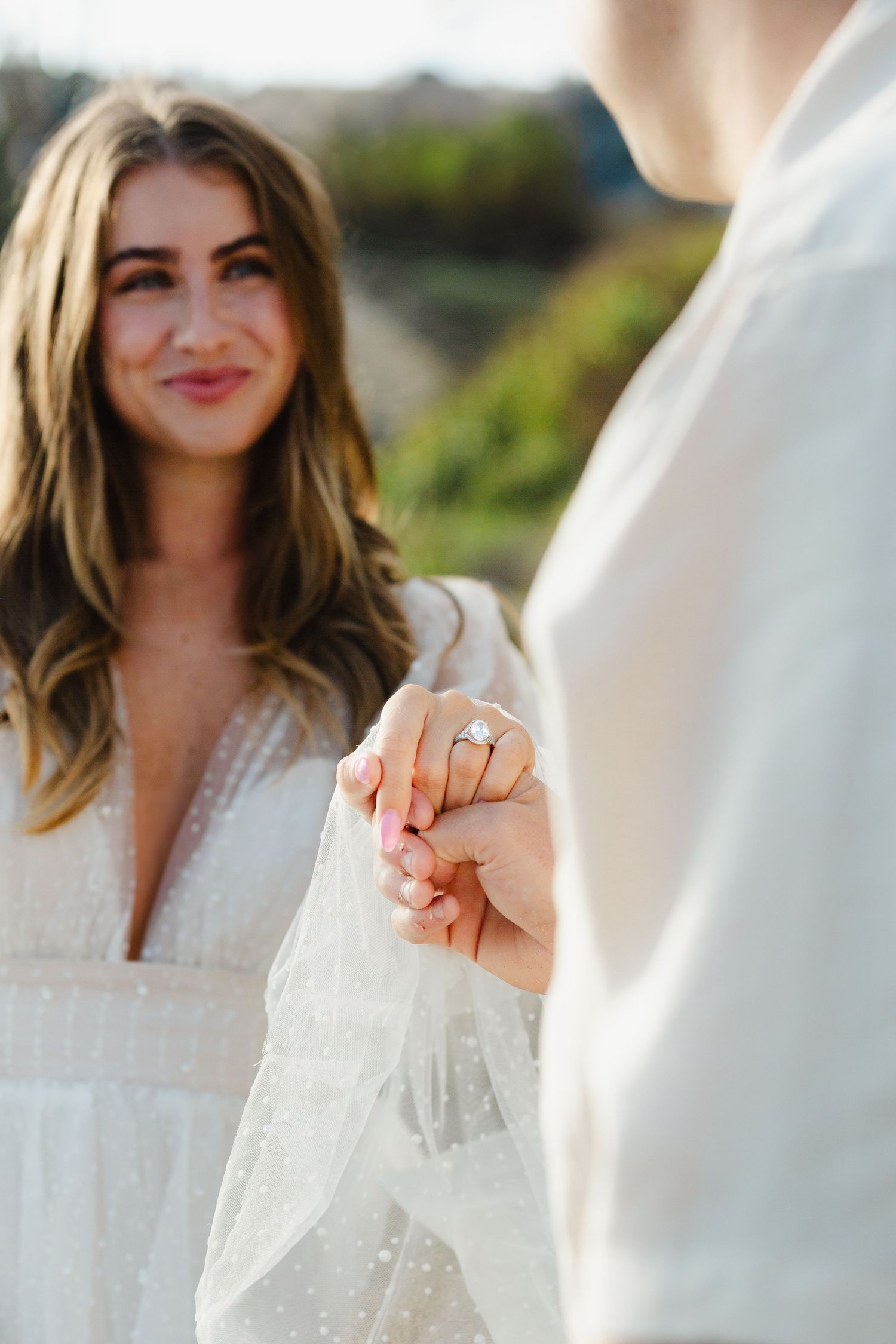 Woman in white lace dress poses outdoors with natural lighting and bokeh background effect.