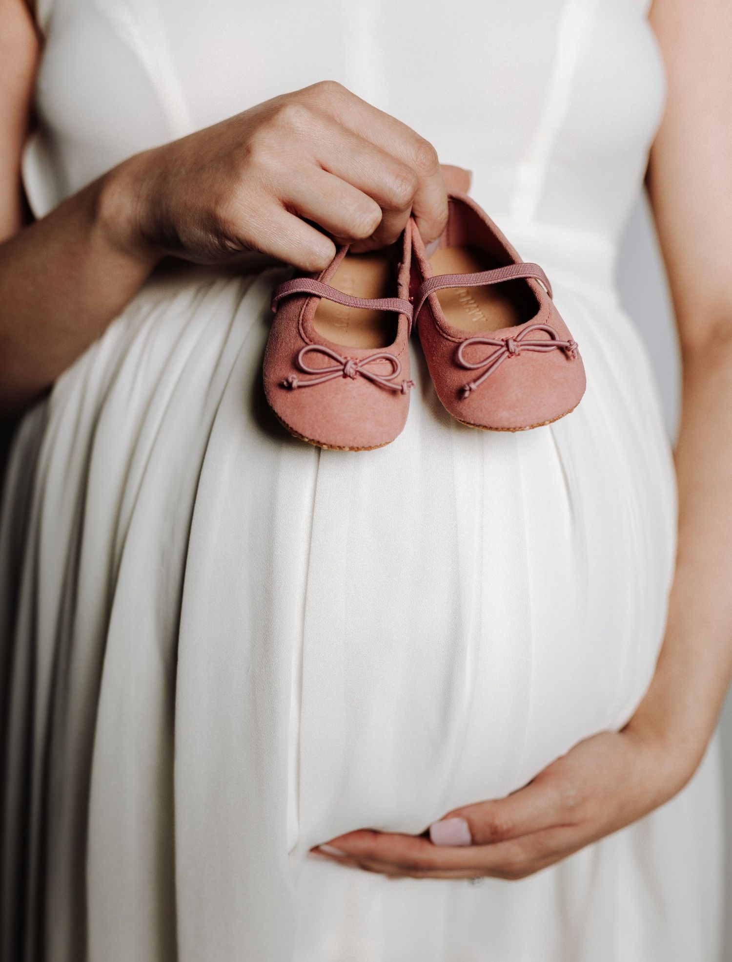 Pregnant woman in white dress holds tiny pink baby shoes against her belly in a tender maternity photo.