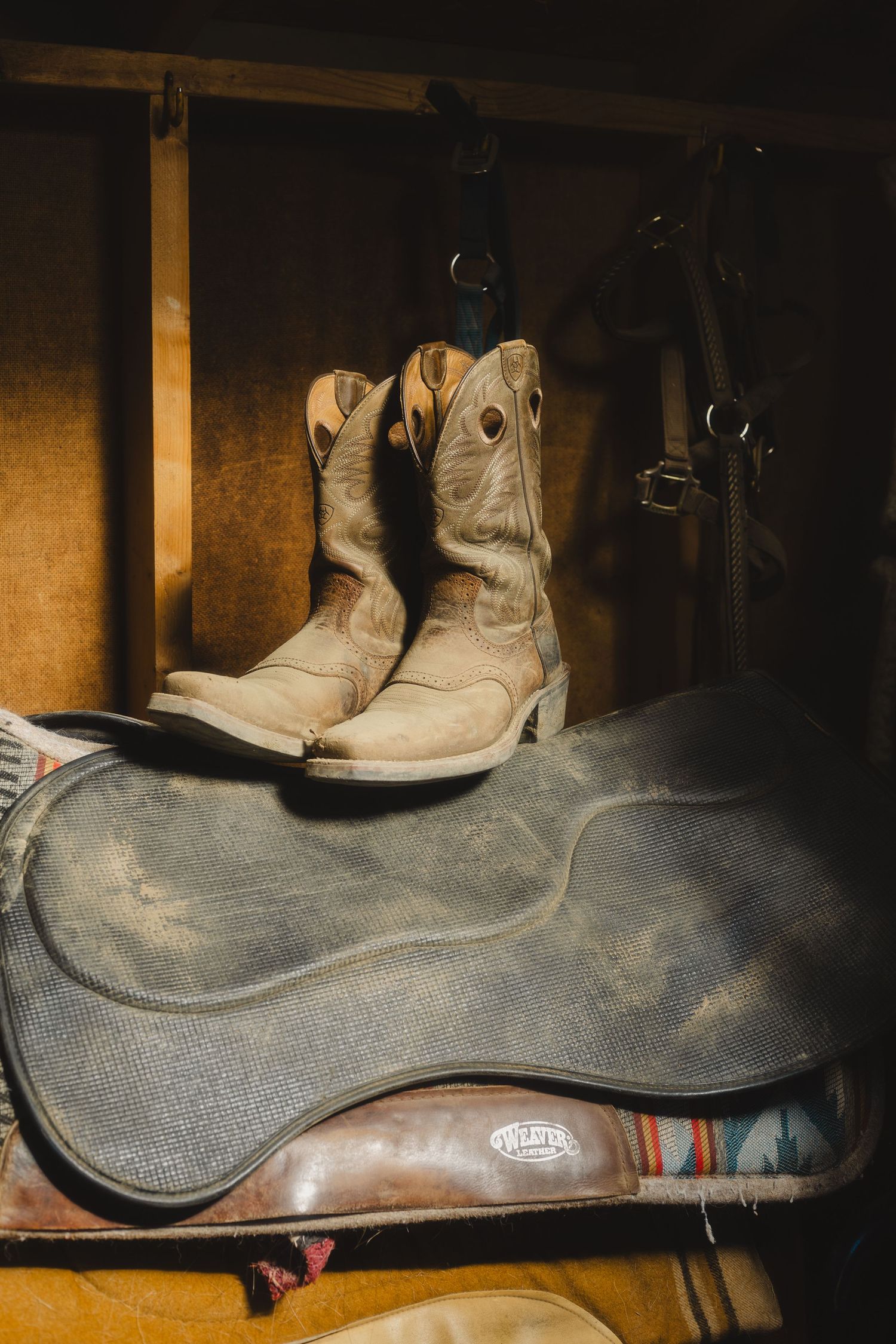 Worn leather cowboy boots sit atop a dusty saddle in dim lighting, creating a rustic western scene.