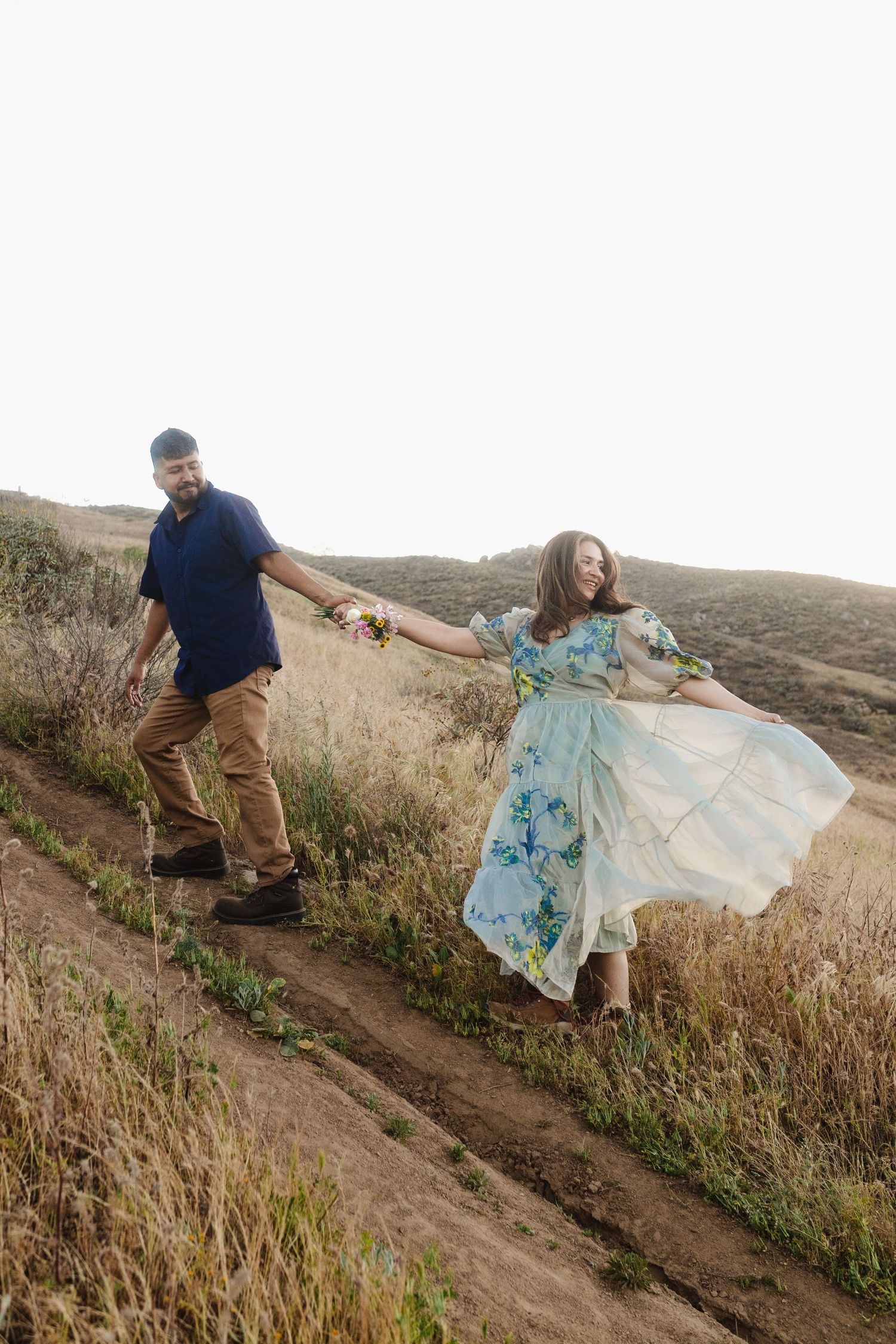 Couple holding hands walking up a hillside trail, woman in floral dress, man in blue shirt, golden grass field backdrop.