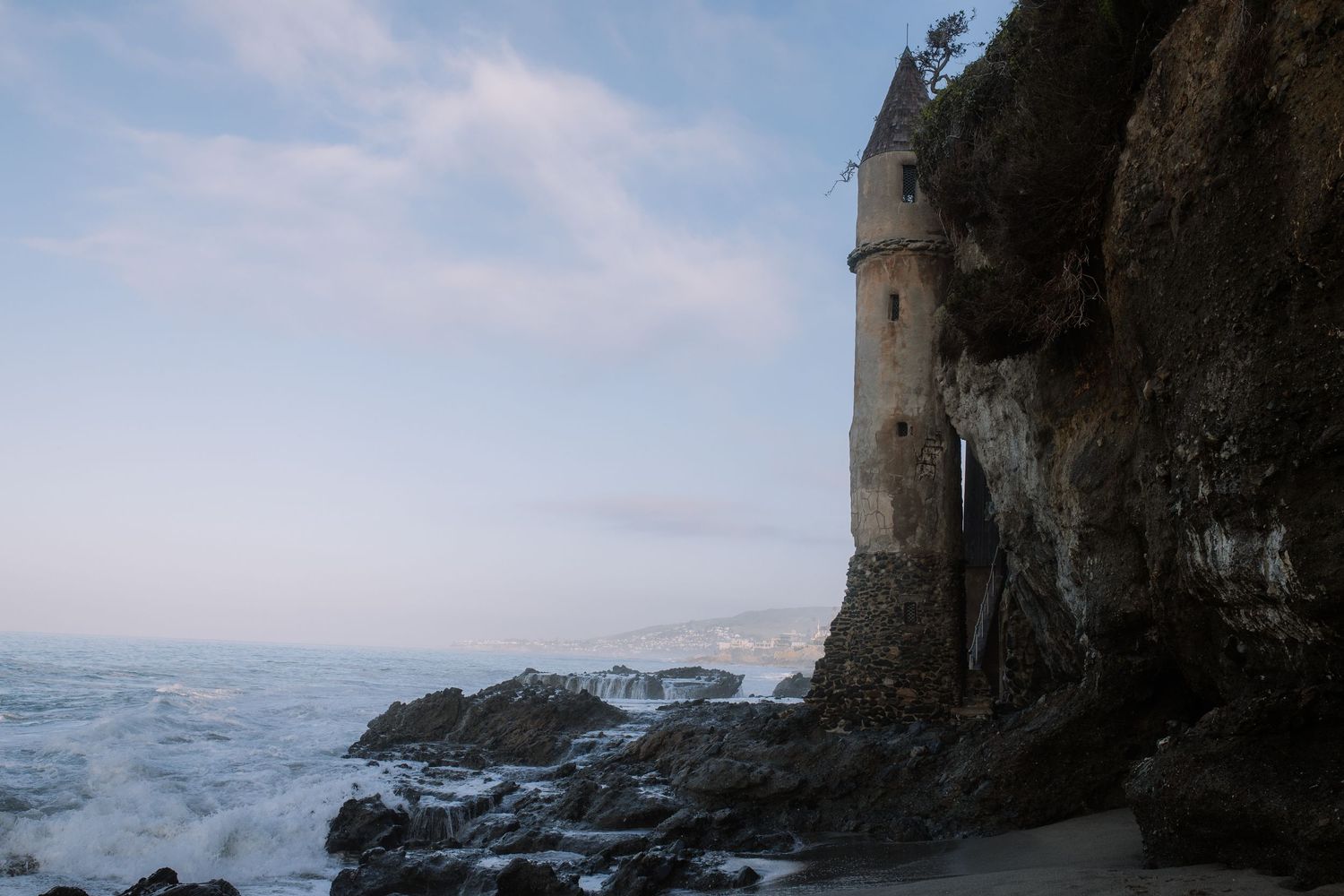 A historic stone tower built into rocky coastal cliffs overlooks crashing ocean waves at dusk.