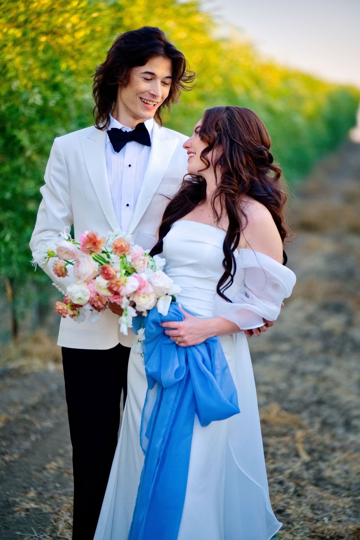 Wedding couple in formal attire with white tuxedo and gown pose together holding bouquet near yellow flowers in nature.