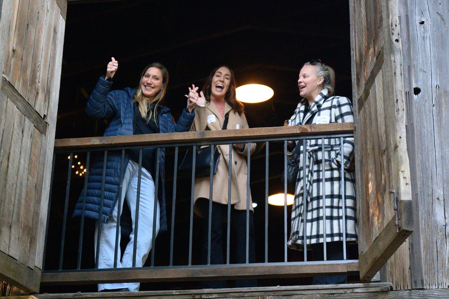 Three people laugh together while standing on a wooden balcony at night with an overhead light illuminating the scene.